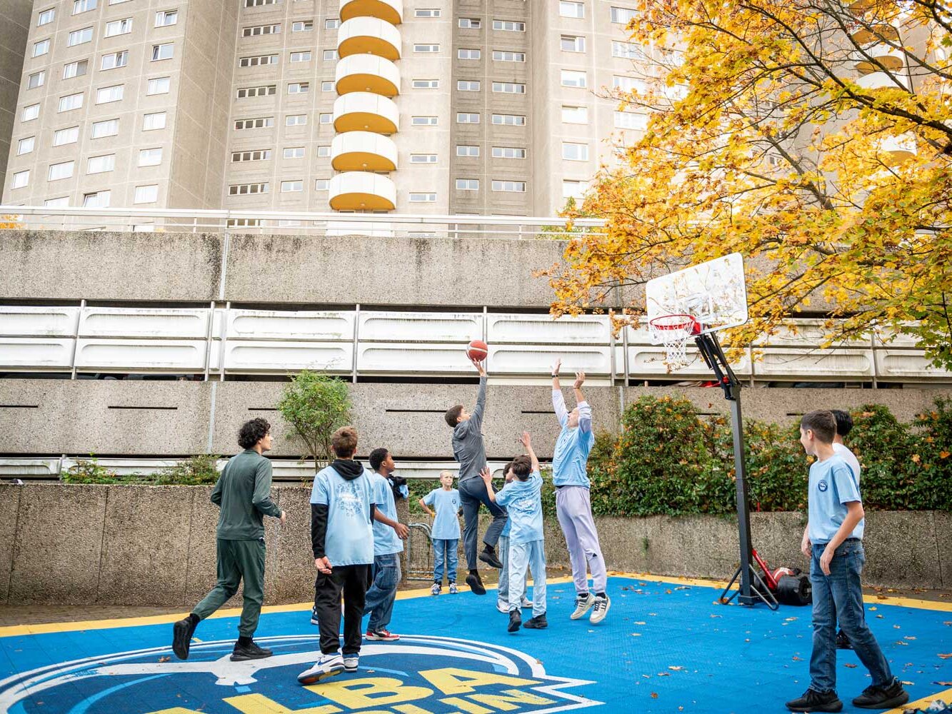 Mehrere Jugendliche spielen Basketball auf einem blau-gelben Outdoor-Court vor einem Wohnhochhaus, während herbstliche Bäume den Platz umrahmen.