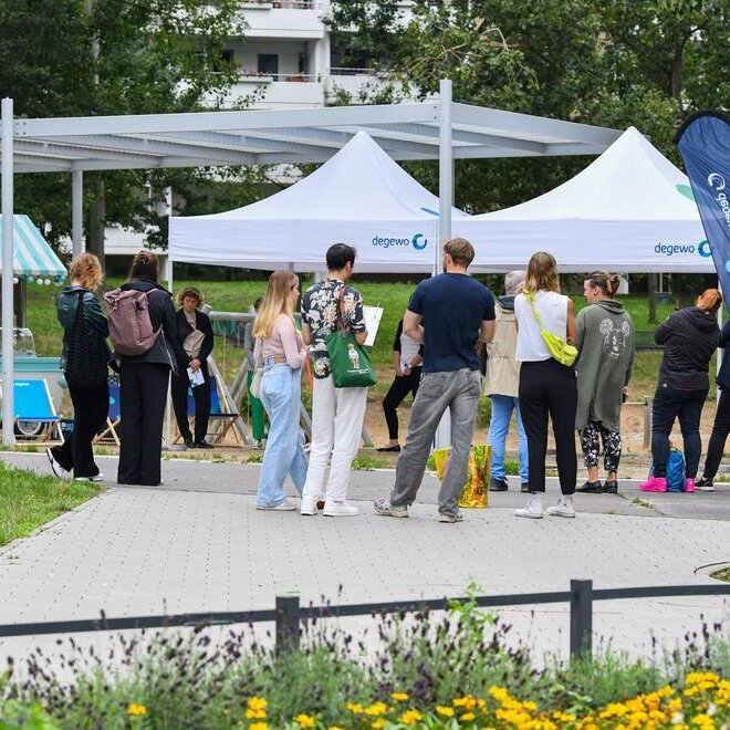 Menschen stehen und gehen auf einem Weg in einer grünen Parkanlage, vor ihnen mehrere weiße Informationszelte mit degewo-Logo; rundherum sind Bäume, Sitzgelegenheiten und bepflanzte Flächen zu sehen.