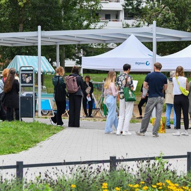 Menschen stehen und gehen auf einem Weg in einer grünen Parkanlage, vor ihnen mehrere weiße Informationszelte mit degewo-Logo; rundherum sind Bäume, Sitzgelegenheiten und bepflanzte Flächen zu sehen.