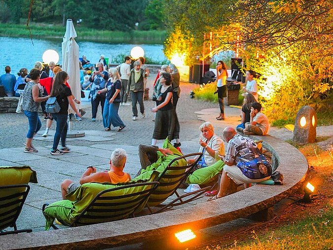 Menschen sitzen an einem See, die Sonne geht unter, die Beleuchtung geht an. Sie unterhalten sich, essen und trinken und genießen die Atmosphäre.