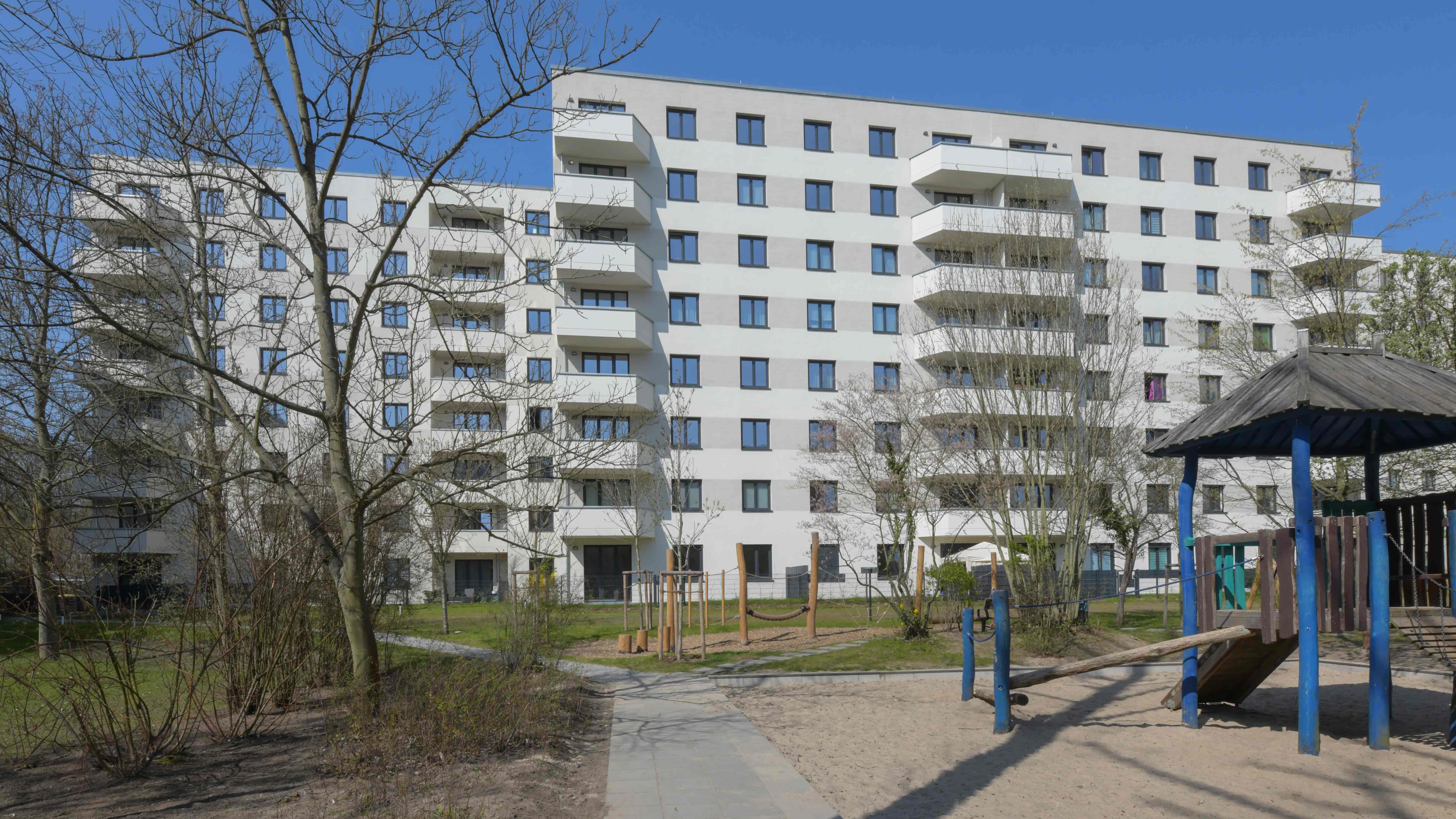 Außenansicht degewo-Neubau Wuhlestraße 2 bis 8 mit Bäumen, Weg und Spielplatz im Vordergrund.