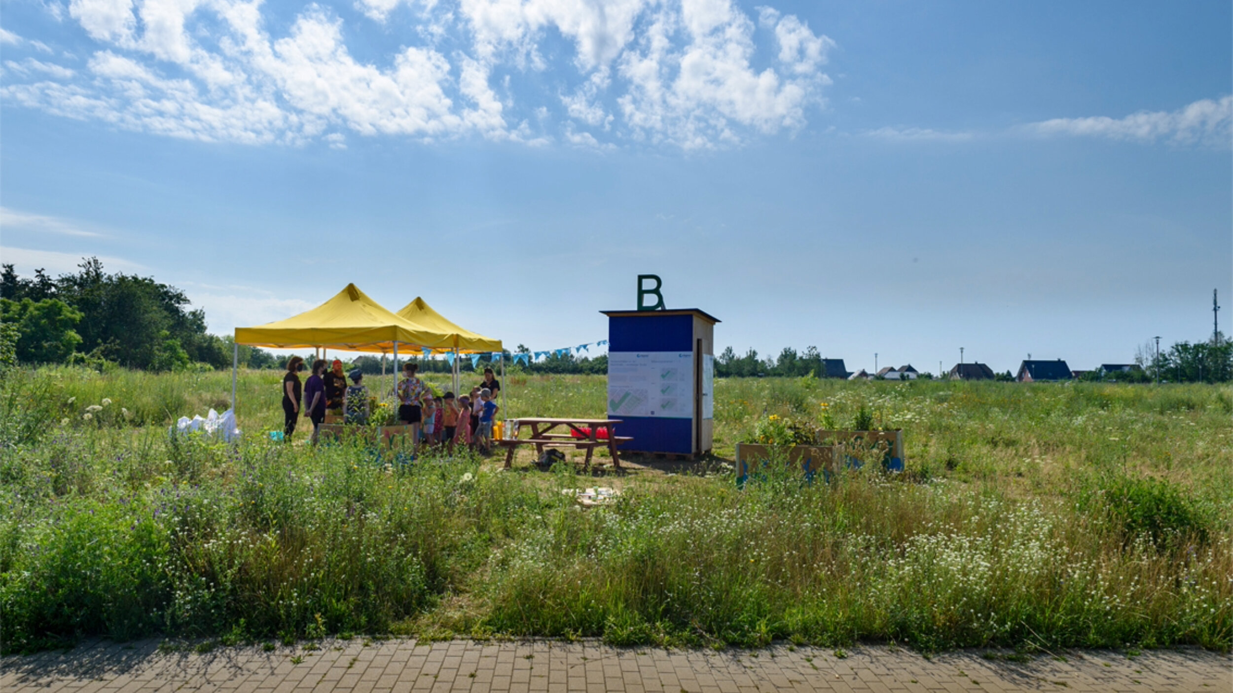 Gesamtansicht einer Beteiligungsveranstaltung auf einer Wiese mit gelben Pavillons, Infotafeln und einer Gruppe von Erwachsenen und Kindern.