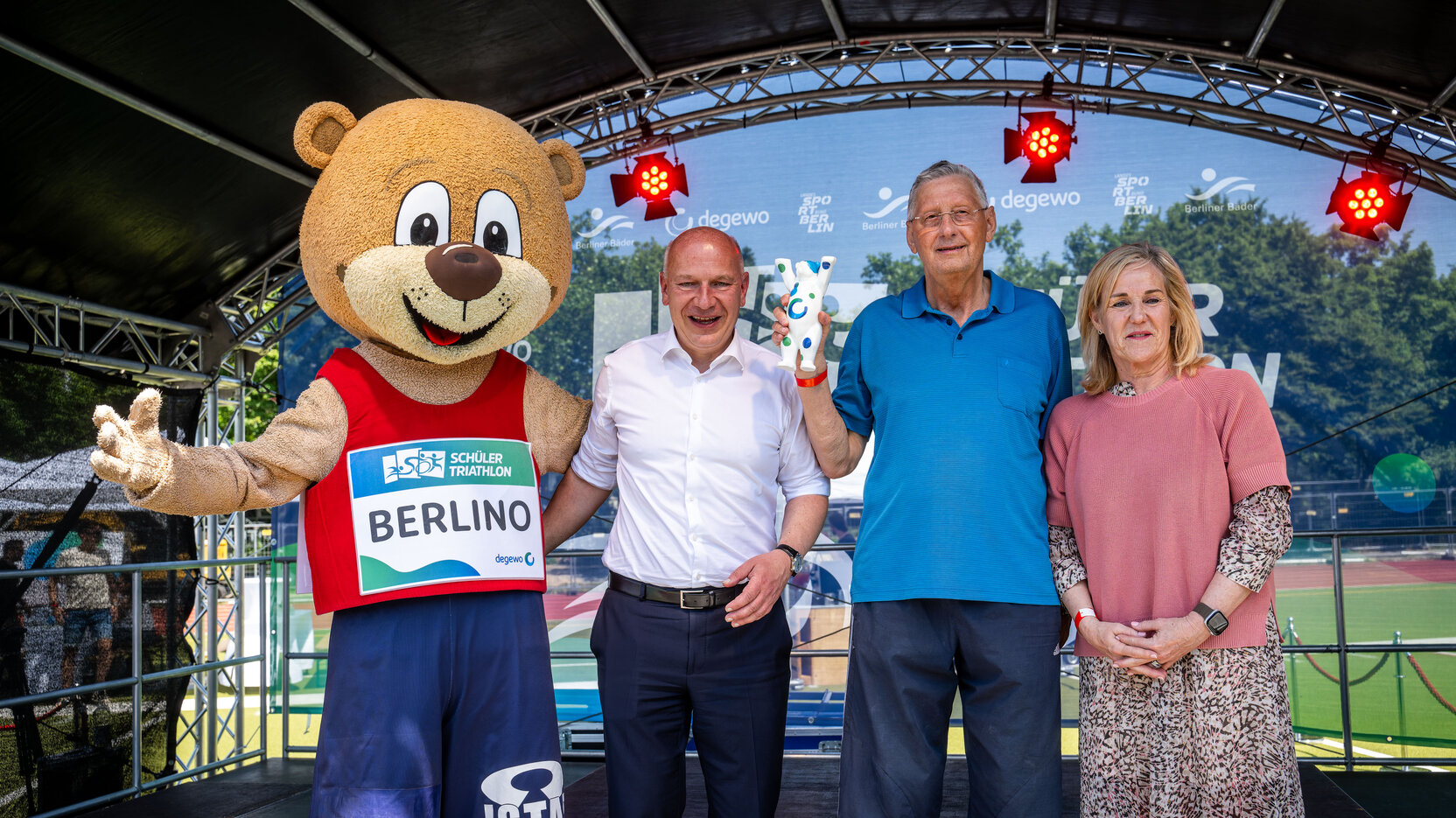 Das Maskottchen Berlino, Berlins Regierender Bürgermeister Kai Wegner, Stanislaw Czaplewski von der Katholischen Grundschule St. Marien und Sandra Wehrmann, Vorstandsmitglied bei degewo, stehen nebeneinander auf einer Bühne.