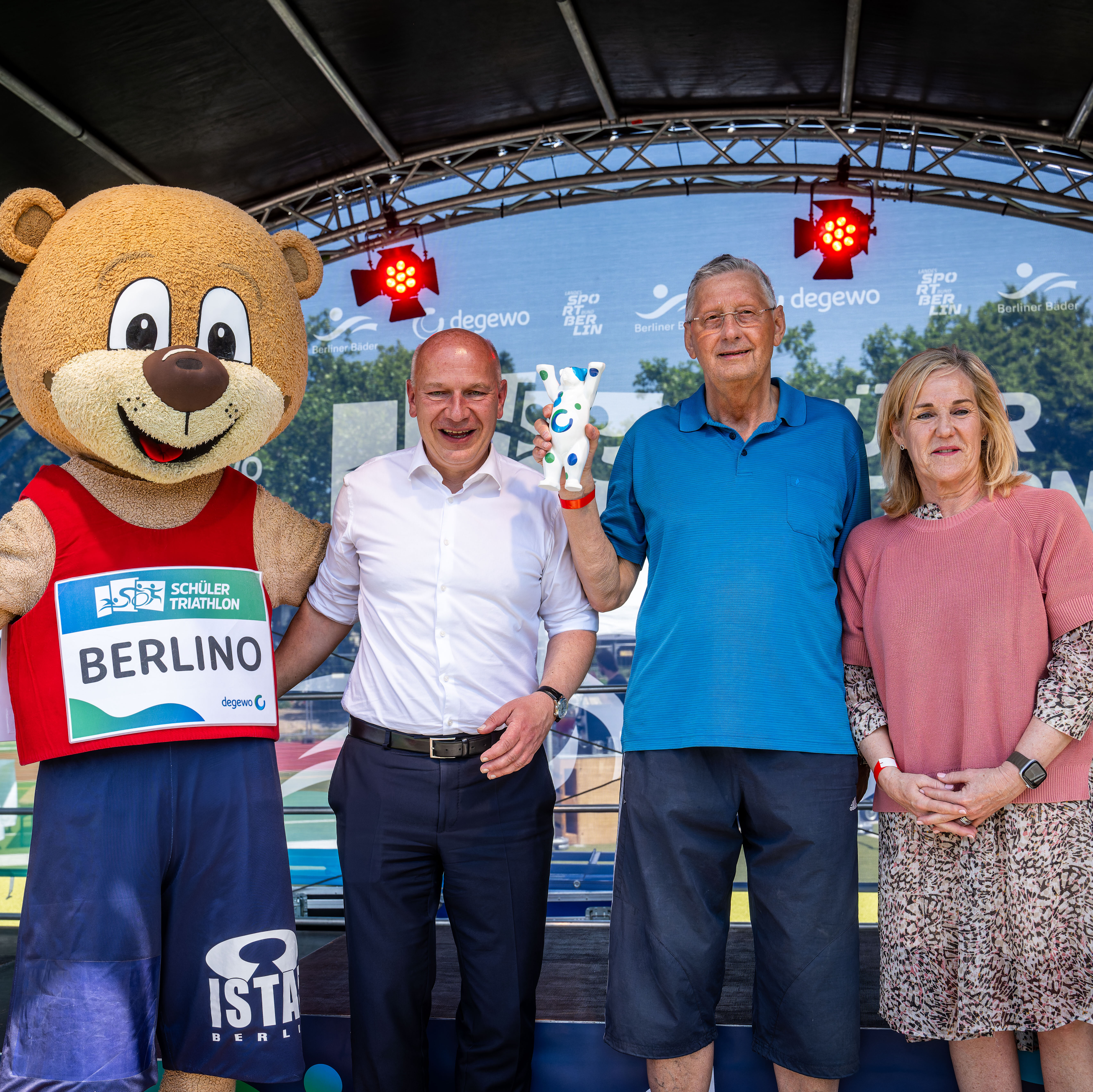 Das Maskottchen Berlino, Berlins Regierender Bürgermeister Kai Wegner, Stanislaw Czaplewski von der Katholischen Grundschule St. Marien und Sandra Wehrmann, Vorstandsmitglied bei degewo, stehen nebeneinander auf einer Bühne.