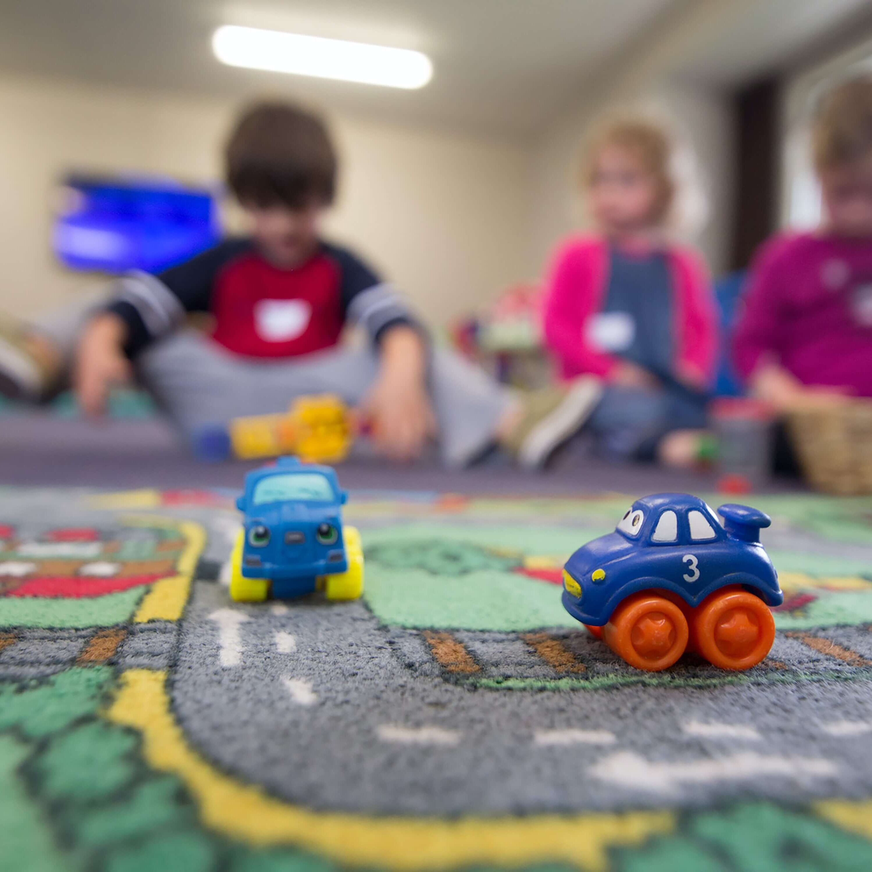 Mehrere Kinder sitzen unscharf im Hintergrund auf dem Boden und spielen, im Vordergrund stehen kleine Spielzeugautos auf einer Spielmatte.