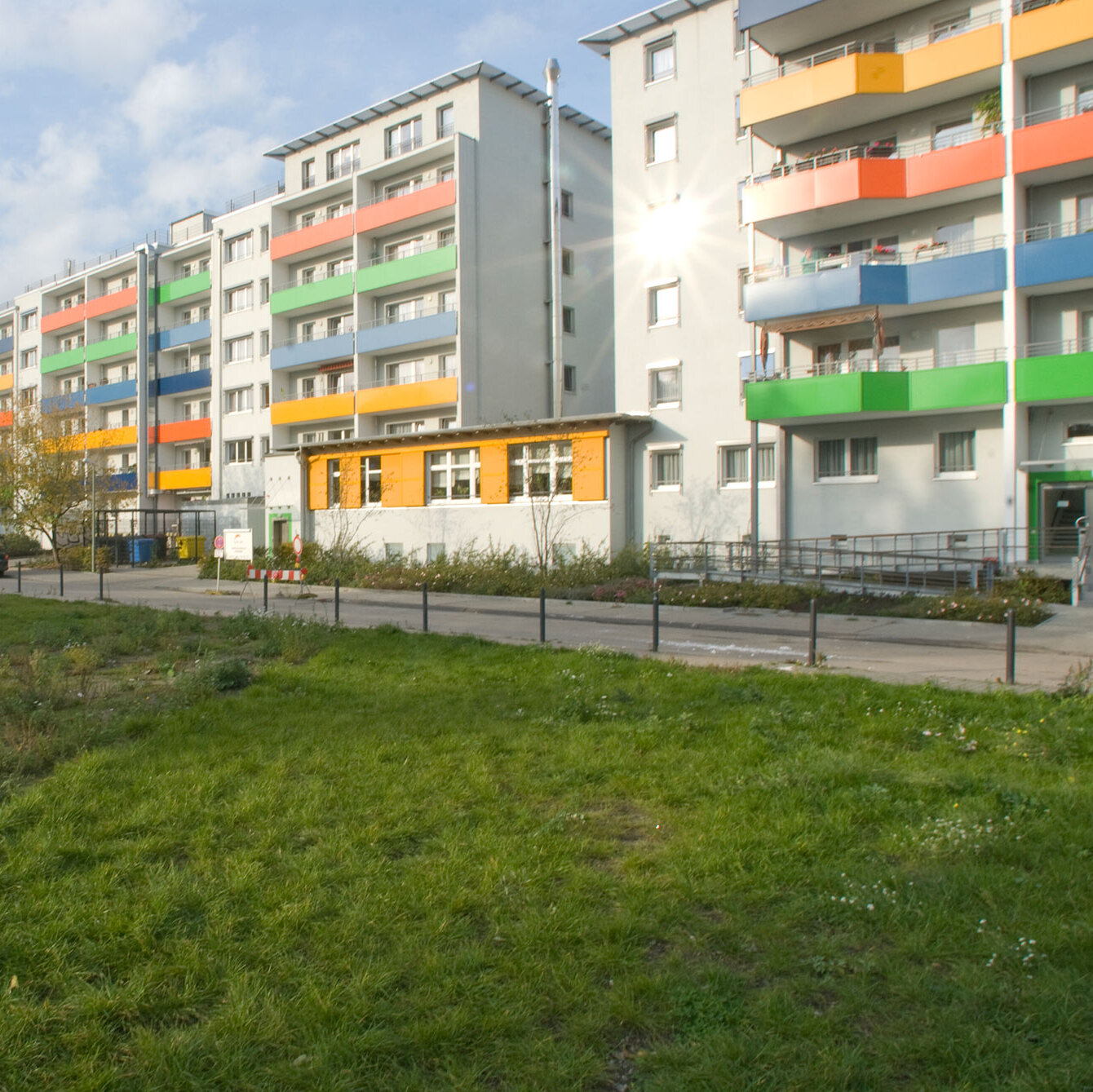 Mehrere modernisierte Wohnhäuser mit farbigen Balkonbrüstungen in Grün, Gelb, Blau und Orange, davor eine Grünfläche und ein Gehweg.