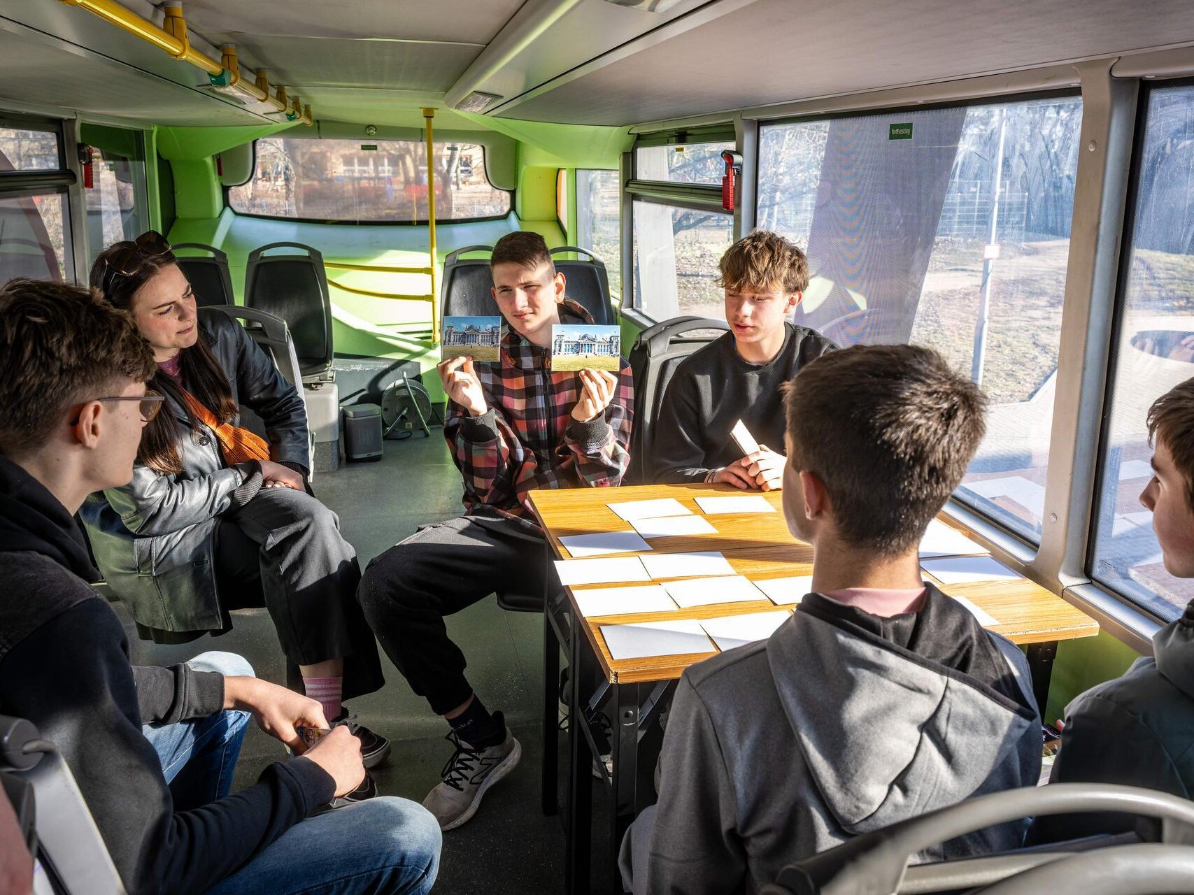Mehrere Jugendliche sitzen um einen Tisch mit umgedeckten Karten. Einer der Jugendlichen hält zwei Karten mit Fotos vom Bundestag nebeneinander.