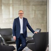 Das Vorstandsmitglied Pascal Atzert steht vor einer Glasscheibe und hat seine Hand auf der Lehne eines Sessels. Er trägt einen Anzug. 