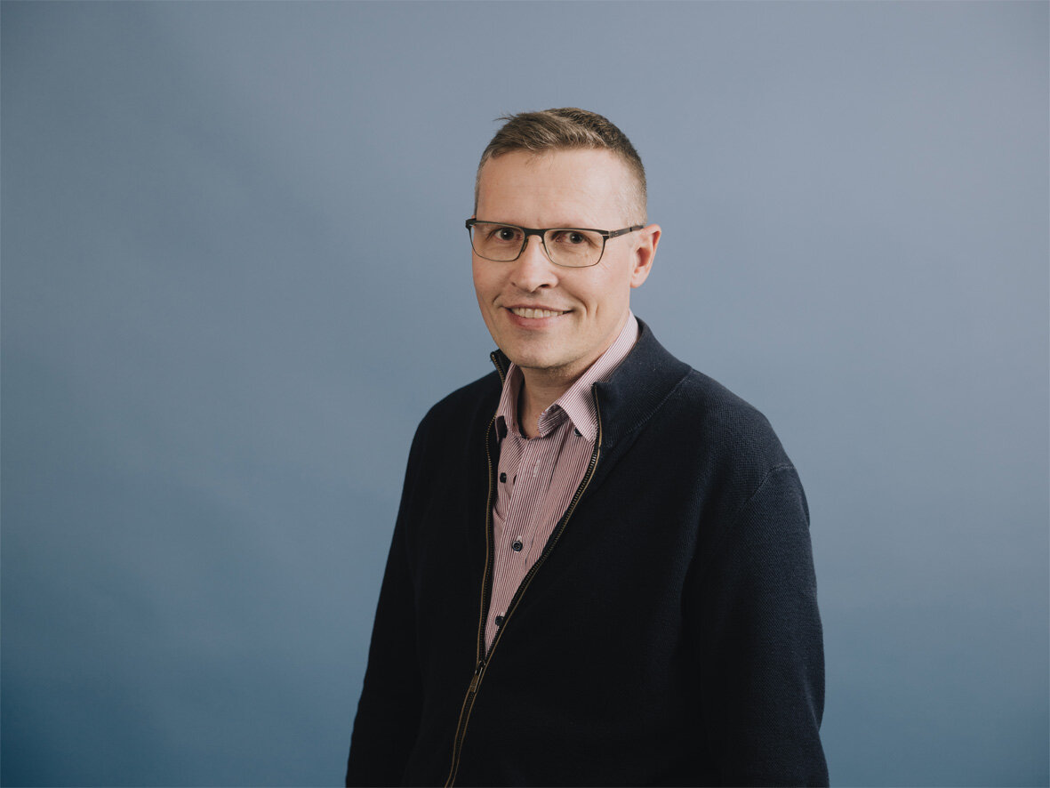 Mann mit Brille trägt eine dunkle Jacke und steht vor einer einfarbigen blauen Wand; er lächelt leicht und blickt direkt in die Kamera.