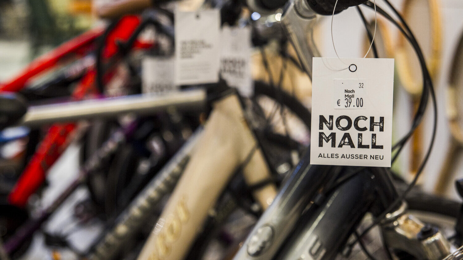 Mehrere gebrauchte Fahrräder stehen dicht aneinandergereiht. Am Lenker eines Rads hängt ein Preisschild mit der Aufschrift „NochMall – Alles außer neu“.
