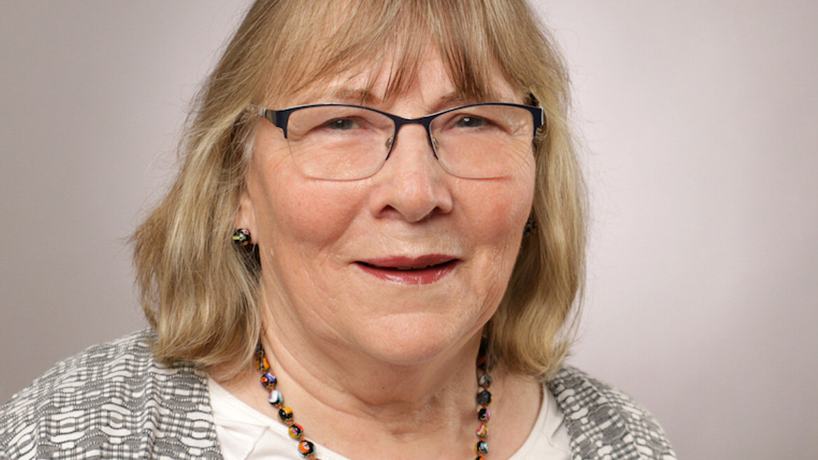 Eine Frau mit blonden, schulterlangen Haaren, Brille, Strickjacke und einer bunten Kette vor hellgrauem Hintergrund.