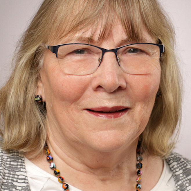Eine Frau mit blonden, schulterlangen Haaren, Brille, Strickjacke und einer bunten Kette vor hellgrauem Hintergrund.