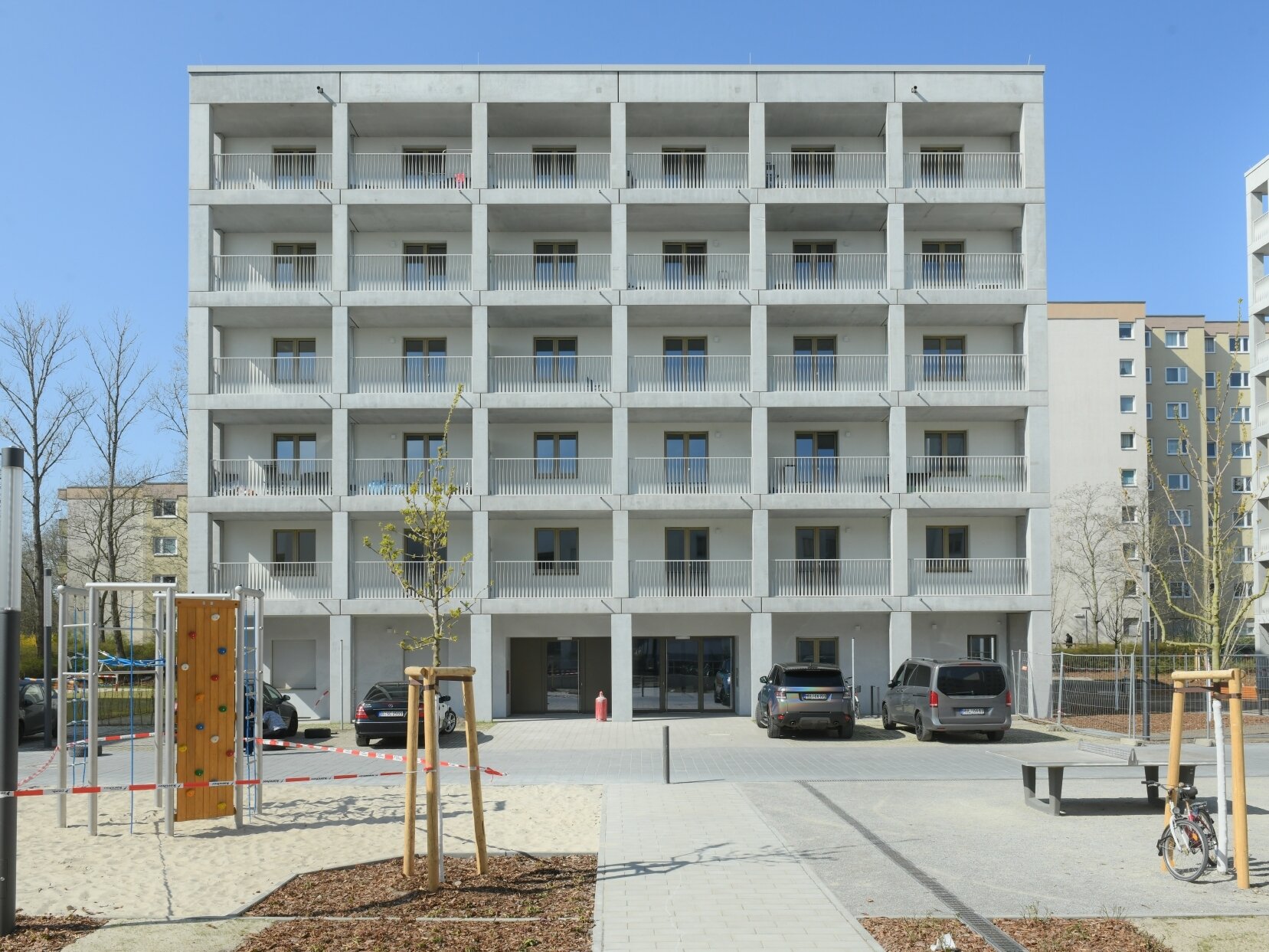 Außenansicht degewo-Neubau Theodor-Loos-Weg 31 bis 35 mit Spielplatz, Straßenlaterne, neu gepflanzten Bäumen, Weg und Tischtennisplatte im Vordergrund.