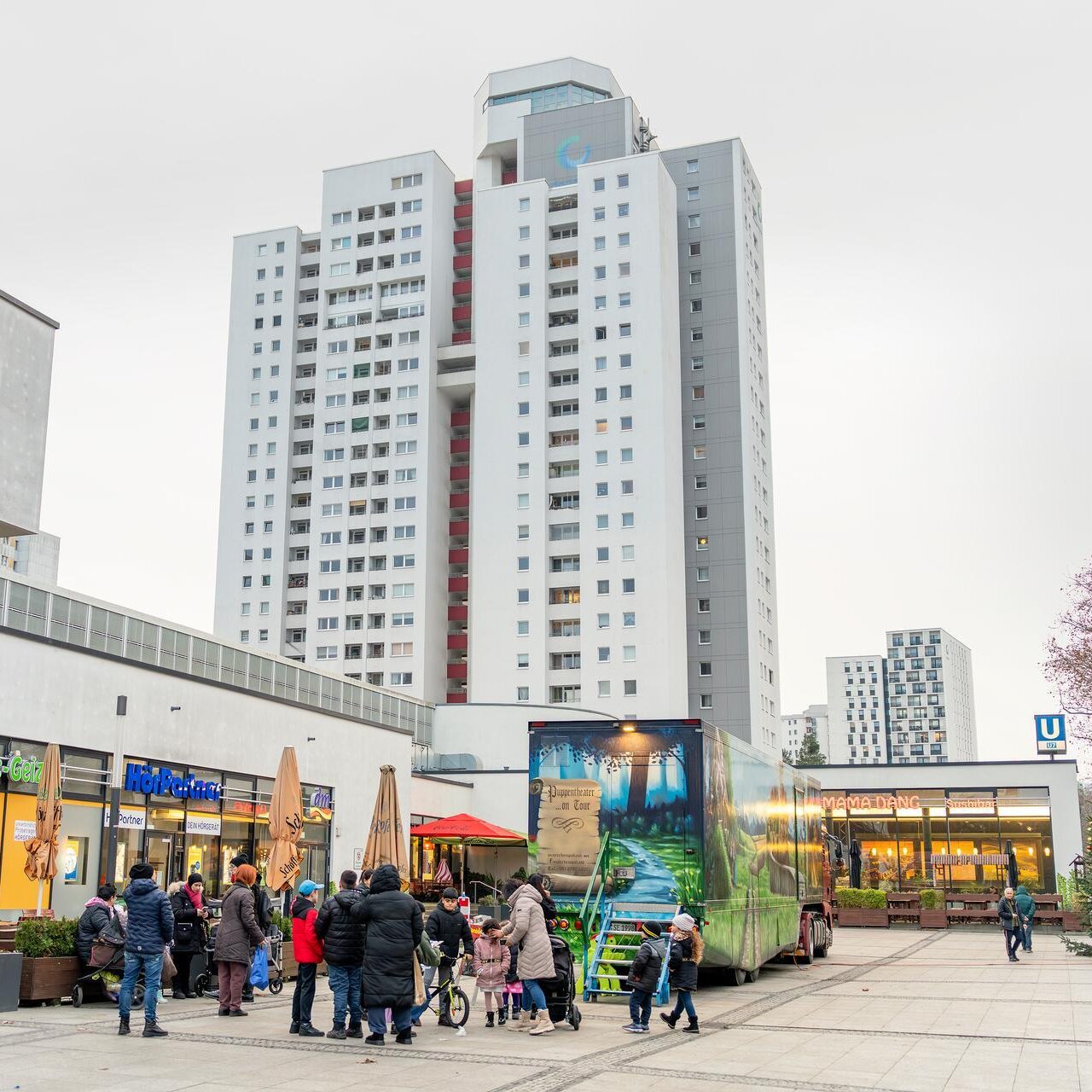 Ansicht des Rotraud-Richter-Platzes mit dem angrenzenden Wutzky-Einkaufscenter. Im Hintergrund ist ein Hochhaus zu sehen. Auf dem Platz stehen Menschen.