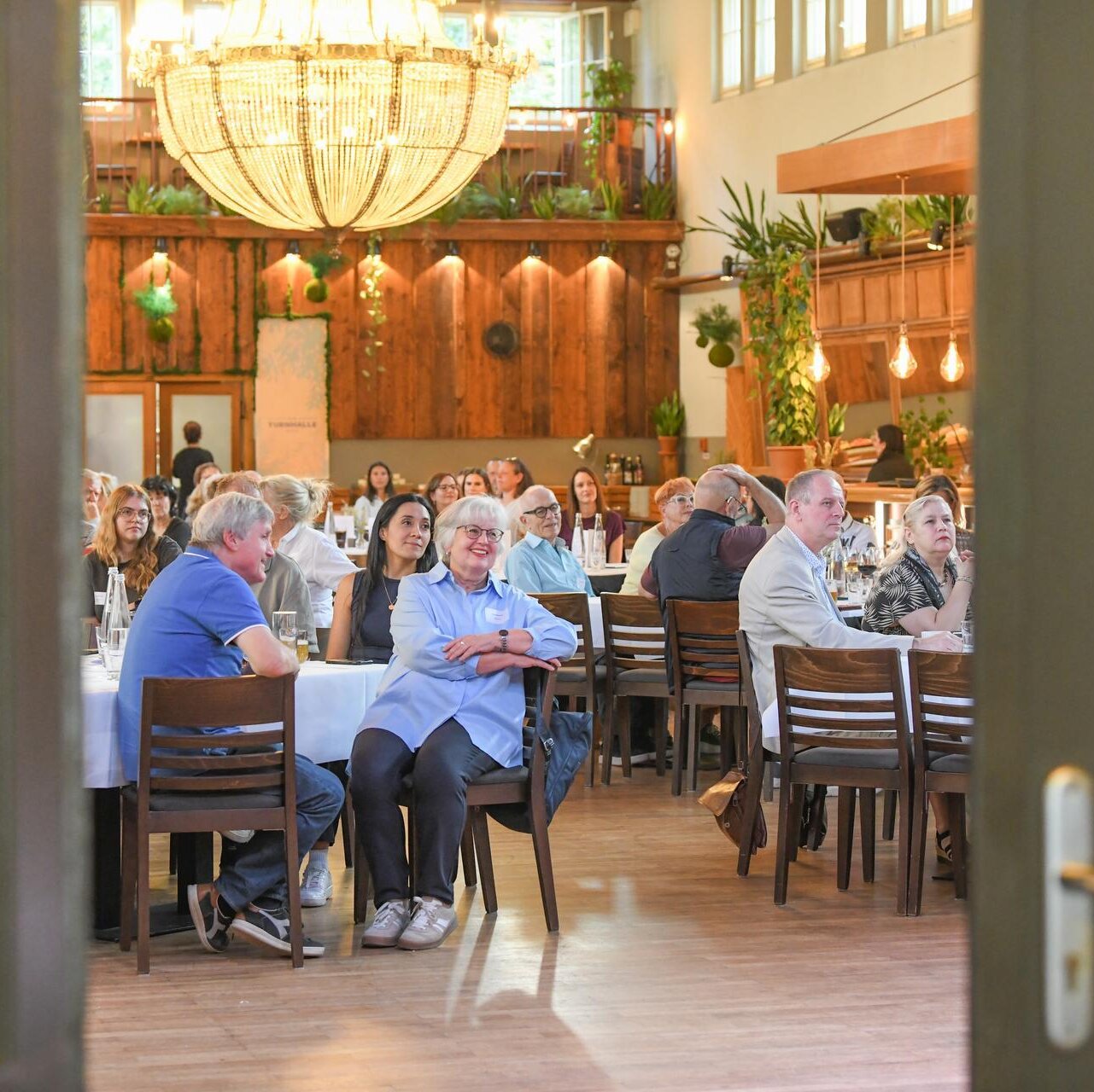 Blick durch eine geöffnete Tür in einen Veranstaltungsraum: Zahlreiche Teilnehmende sitzen an Tischen und verfolgen eine Veranstaltung. Ein großer Kronleuchter hängt zentral im Raum, Holzverkleidungen und Pflanzen prägen das Ambiente.