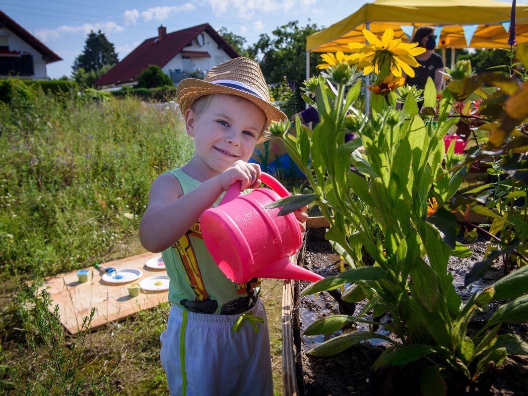 Ein Kind gießt mit einer pinken Gießkanne Pflanzen in einem Hochbeet. Die Aktion ist Teil eines kreativen Beteiligungsangebots im Freien.