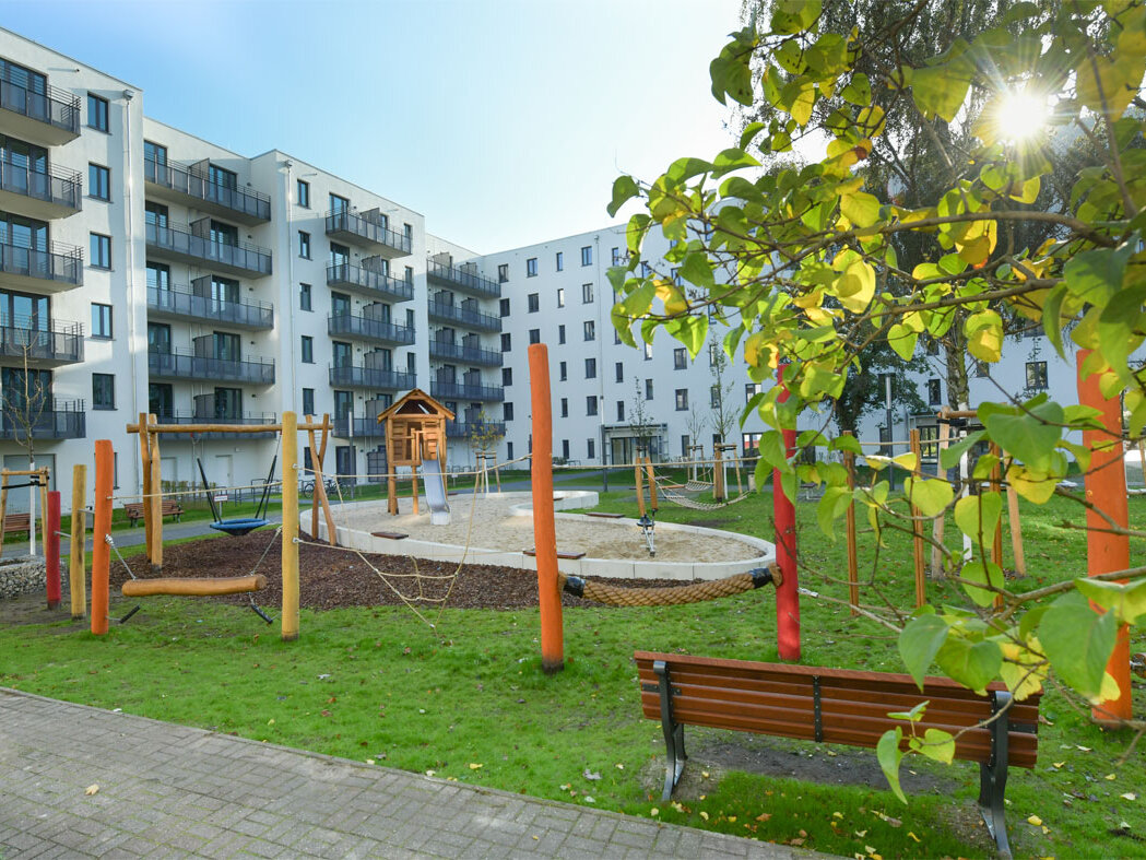 Außenansicht degewo-Neubau Mahlower Straße 2 bis 4 A mit Weg, Sitzbank, Grünfläche, Spielplatz und beblätterten Baumzweigen rechts im Vordergrund.