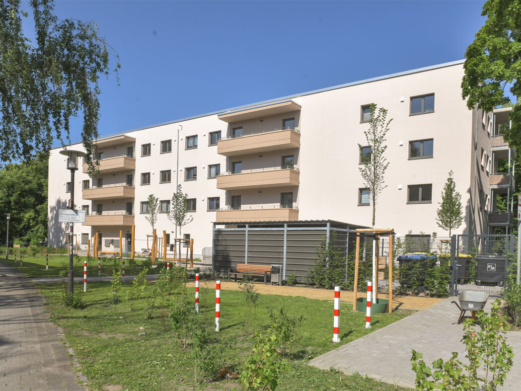 Außenansicht degewo-Neubau Ottomar-Geschke-Straße mit Wegen, Grünflächen, neu gepflanzten Bäumen und Straßenlaterne im Vordergrund.