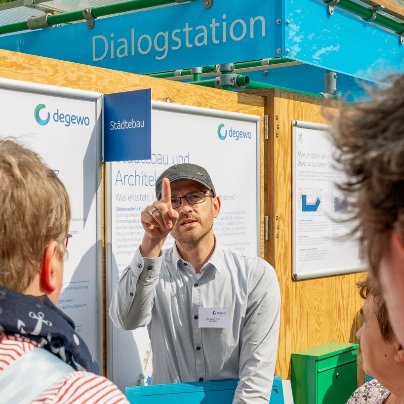 degewo-Mitarbeiter im Gespräch mit Anwohnenden an der Dialogstation, erläutert Planungsinhalte anhand ausgestellter Informationstafeln.