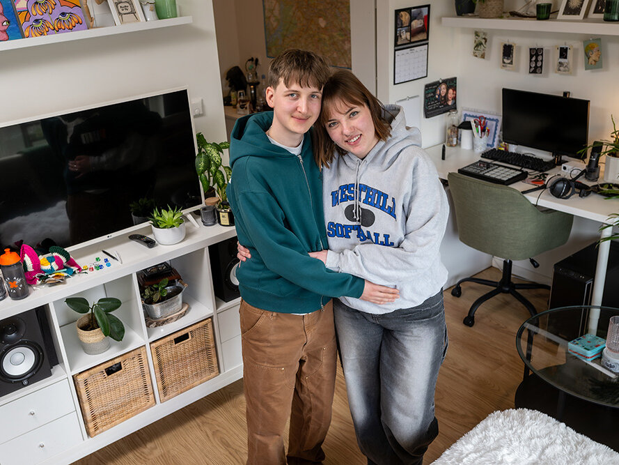 Ein Paar steht umarmt in einem hell eingerichteten Wohnzimmer mit Regalen, Schreibtisch und Fernseher. Beide lächeln in die Kamera.