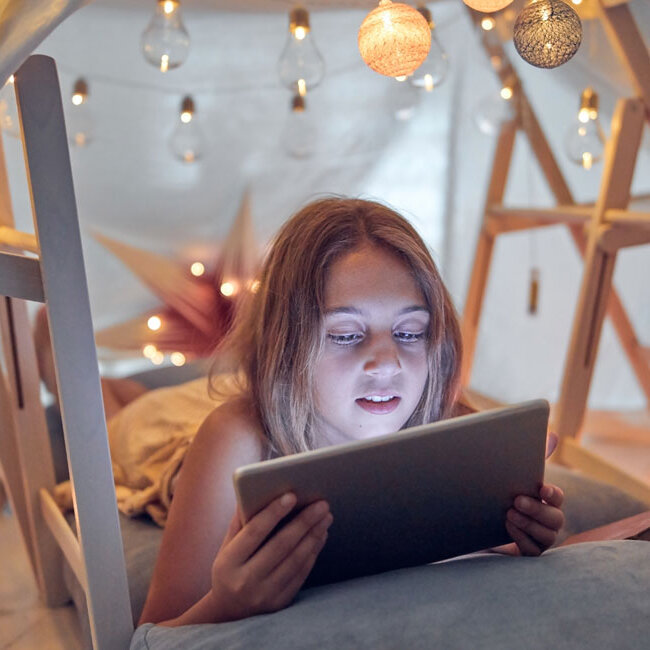 Kind liegt in einem selbstgebauten Zelt aus Decken und Holzgestellen und schaut auf ein Tablet, umgeben von warm leuchtenden Lichterketten und brennenden Kerzen.