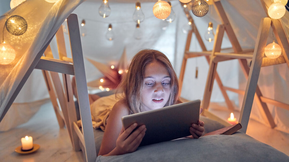 Kind liegt in einem selbstgebauten Zelt aus Decken und Holzgestellen und schaut auf ein Tablet, umgeben von warm leuchtenden Lichterketten und brennenden Kerzen.