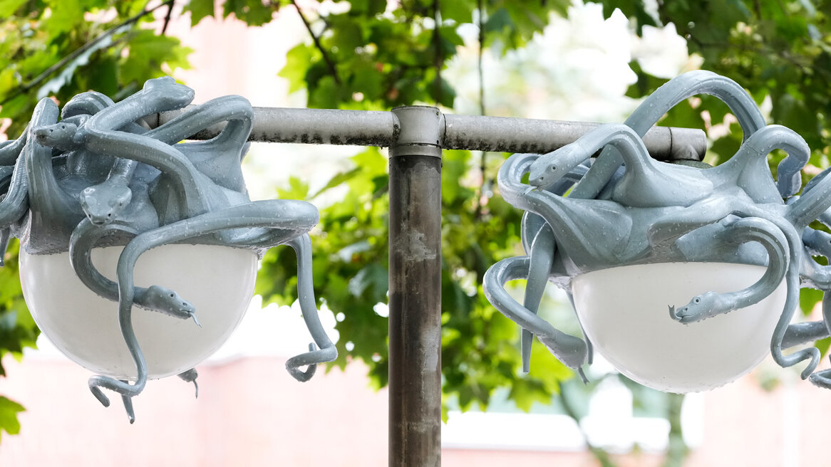 Zwei Straßenlampen, deren kugelförmige Leuchten von zahlreichen grauen, schlangenartigen Skulpturen umschlungen sind, vor grünem Blätterdach.