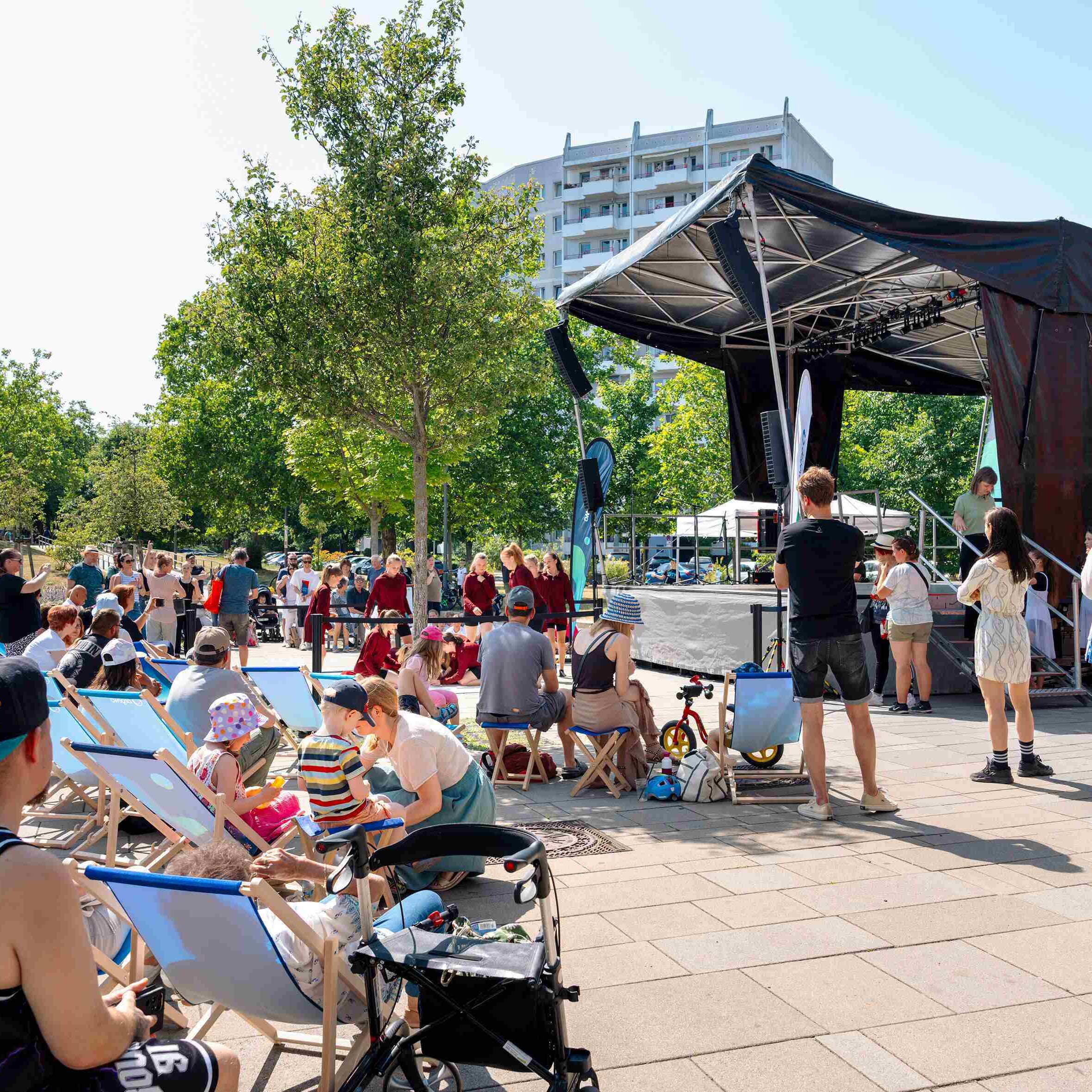 Vor einer Open-Air-Bühne sitzt das Publikum auf Klappstühlen. Die Sonne scheint. 