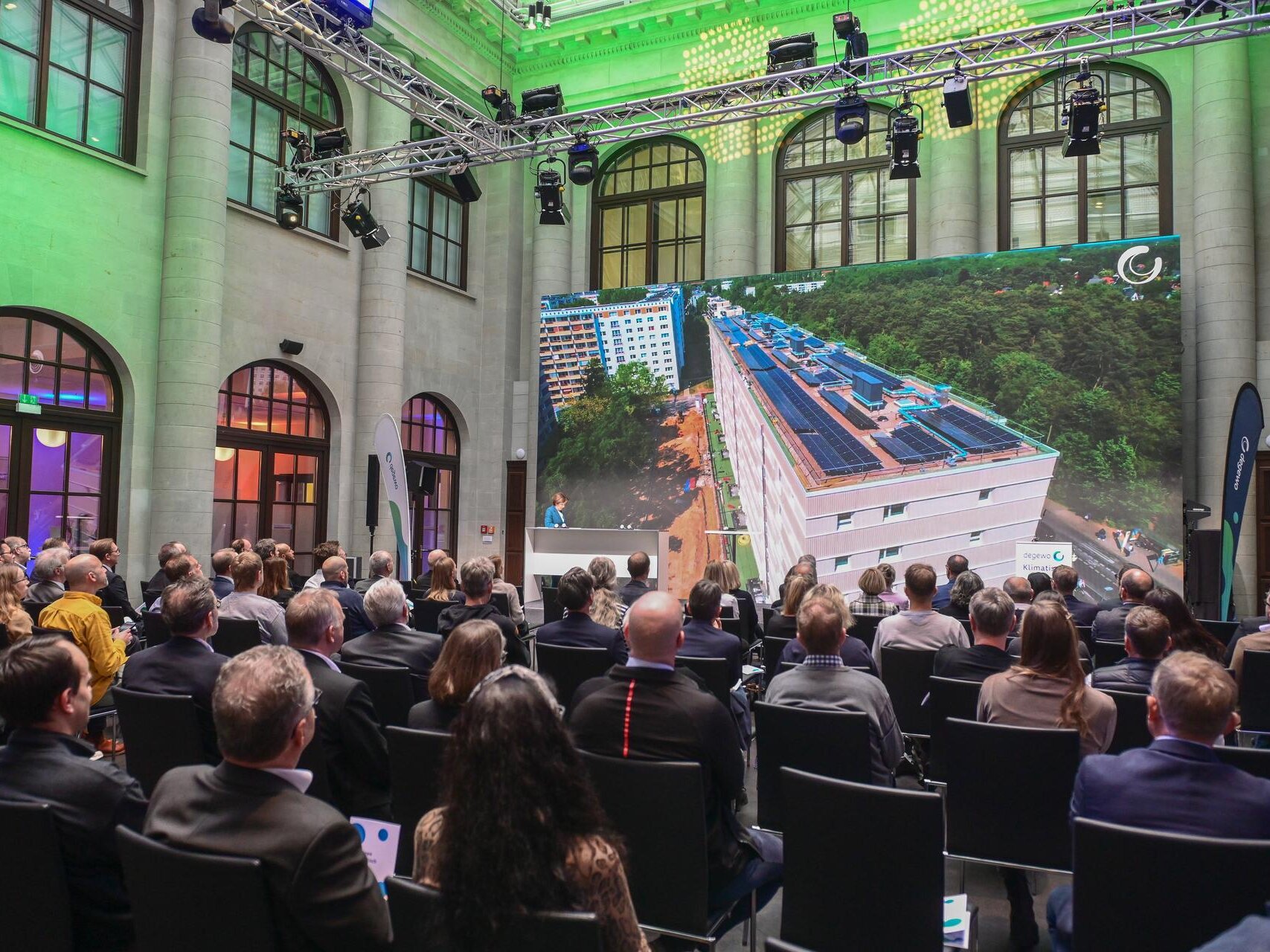 Teilnehmende einer Fachveranstaltung sitzen in einer großen Halle und verfolgen eine Präsentation. Auf der großformatigen Leinwand wird ein Wohngebäude mit Photovoltaikanlagen auf dem Dach gezeigt.