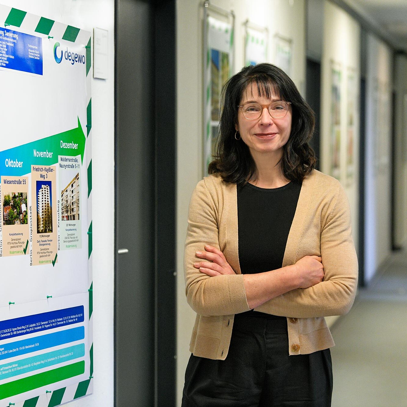 Frau mit Brille und verschränkten Armen steht in einem Büroflur neben einer großen Infotafel mit Zeitplan und Projektübersicht der degewo und blickt in die Kamera.
