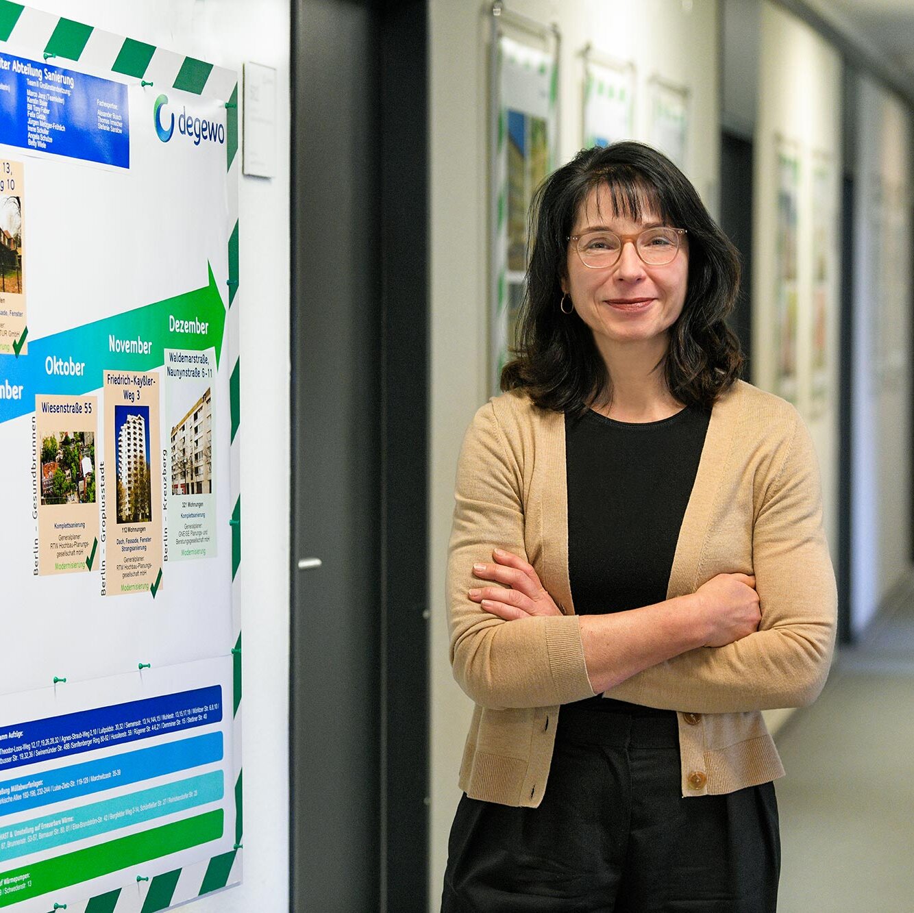Frau mit Brille und verschränkten Armen steht in einem Büroflur neben einer großen Infotafel mit Zeitplan und Projektübersicht der degewo und blickt in die Kamera.