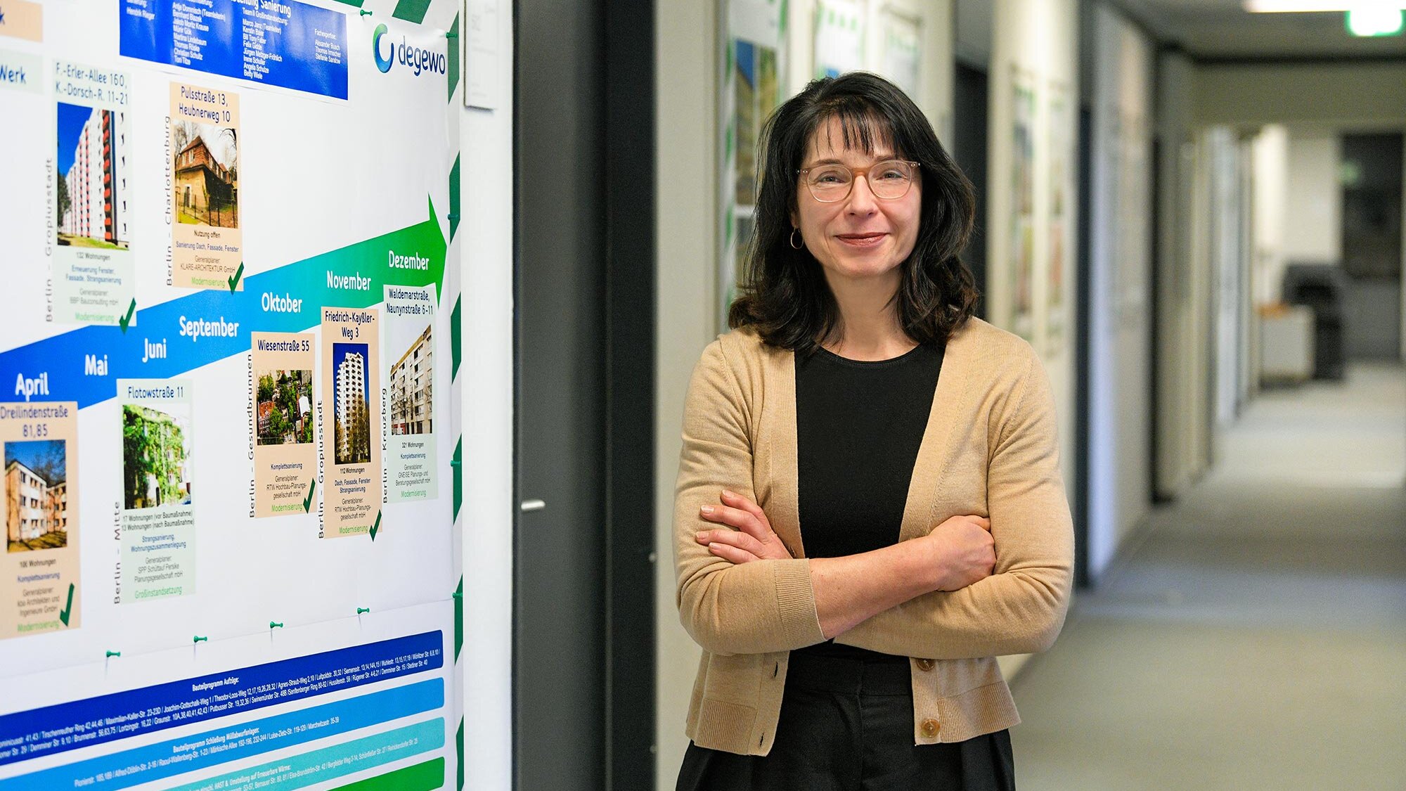 Frau mit Brille und verschränkten Armen steht in einem Büroflur neben einer großen Infotafel mit Zeitplan und Projektübersicht der degewo und blickt in die Kamera.