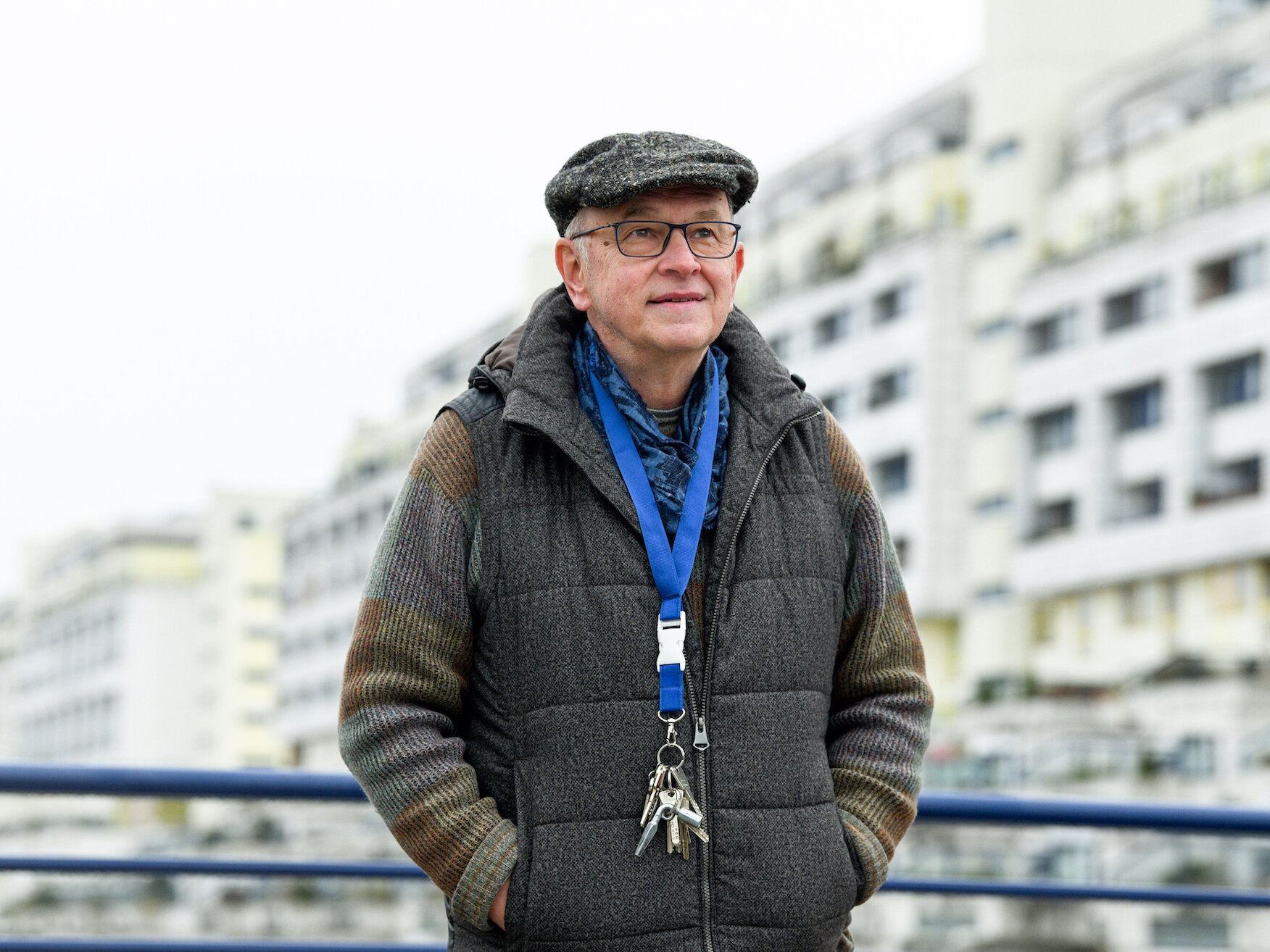 Mann mit Mütze und Brille steht auf einem Balkon mit Blick auf ein großes Wohngebäude, trägt eine Weste und einen Schlüsselbund am Band um den Hals.
