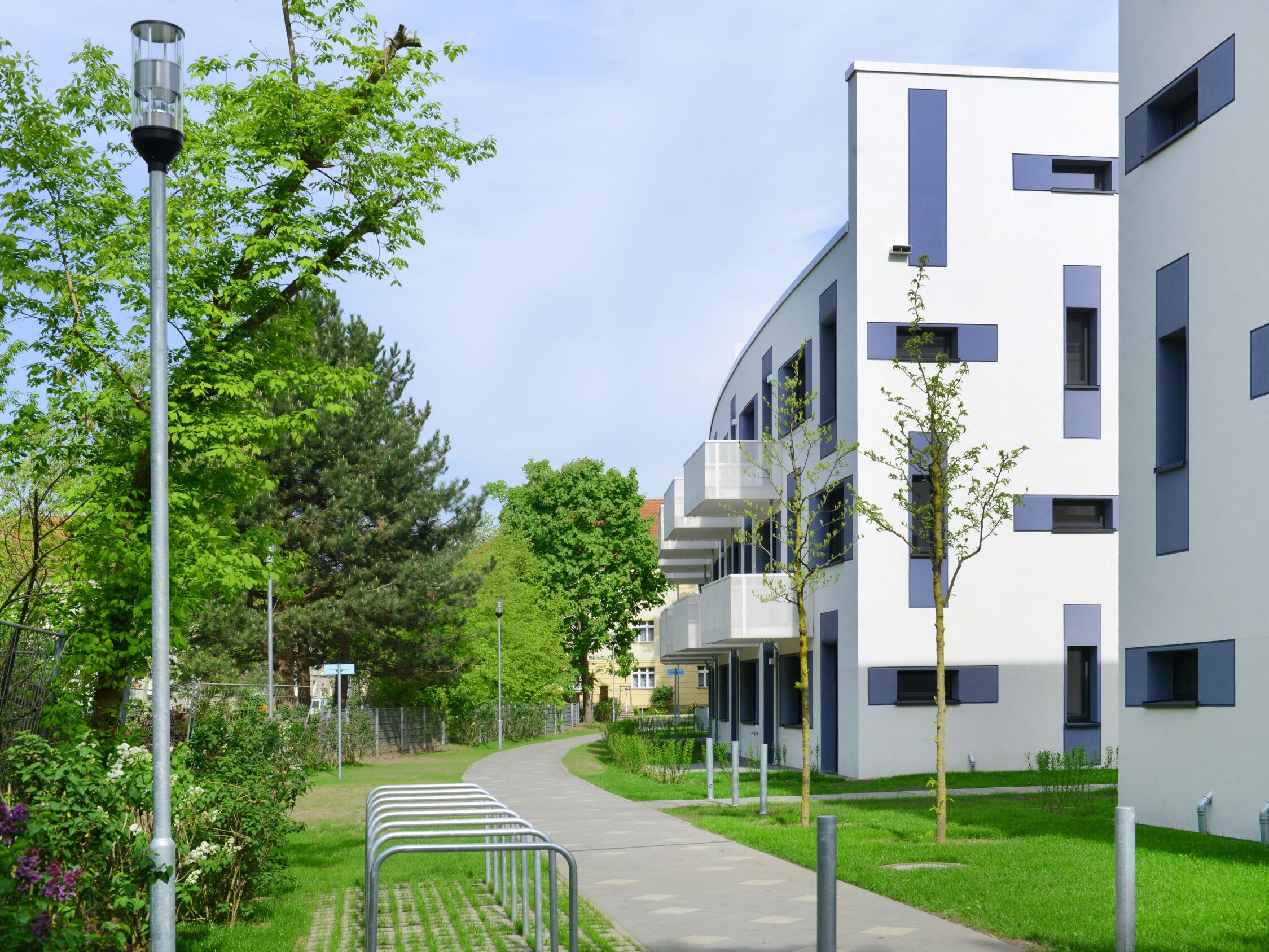 Außenansicht degewo-Neubau Joachimstraße 8 A bis E mit Fliederbüschen, Straßenlaterne, Fahrradständern, Weg und Rasenfläche im Vordergrund.