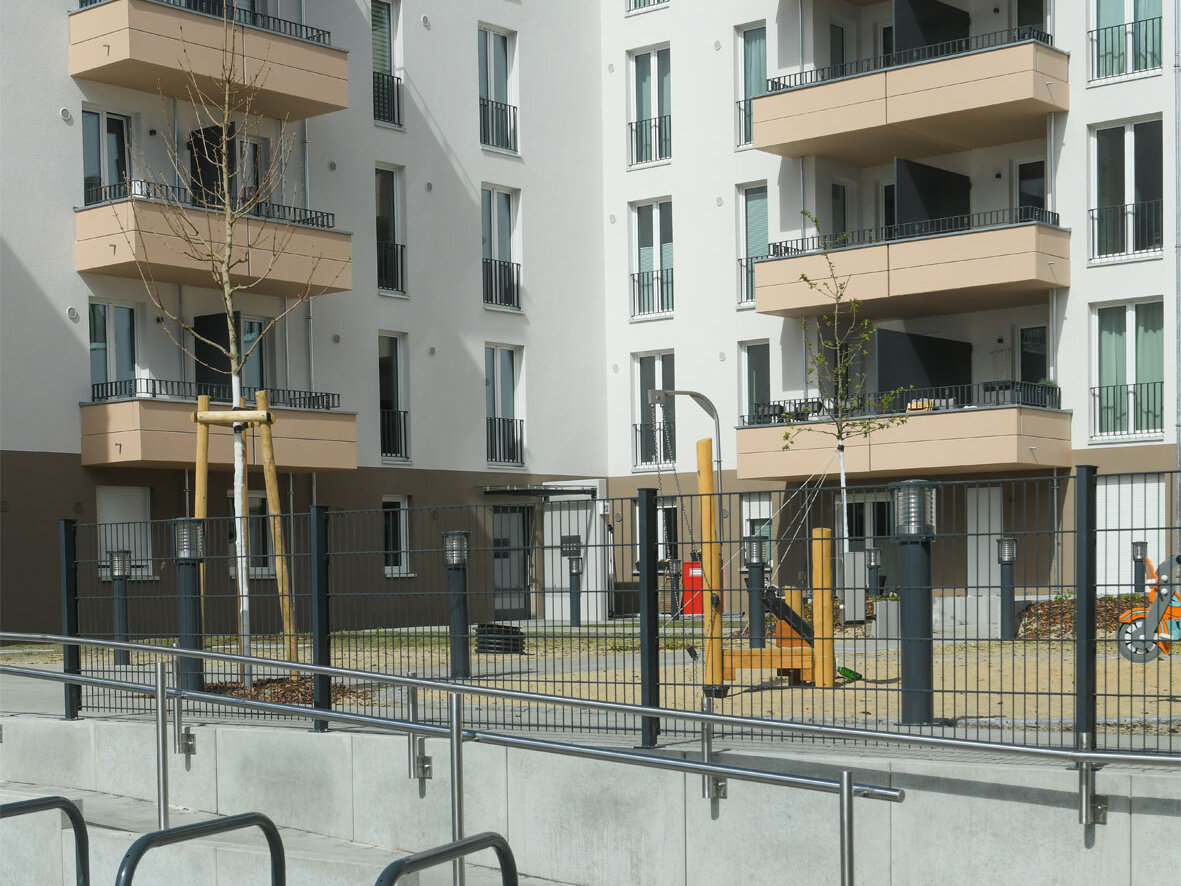 Außenansicht degewo-Neubau Schwartzkopffstraße 20 bis 28: Hofseite mit umzäuntem Spielplatz. Im Vordergrund: gepflasterter Weg mit Geländer und drei Fahrradständer links.