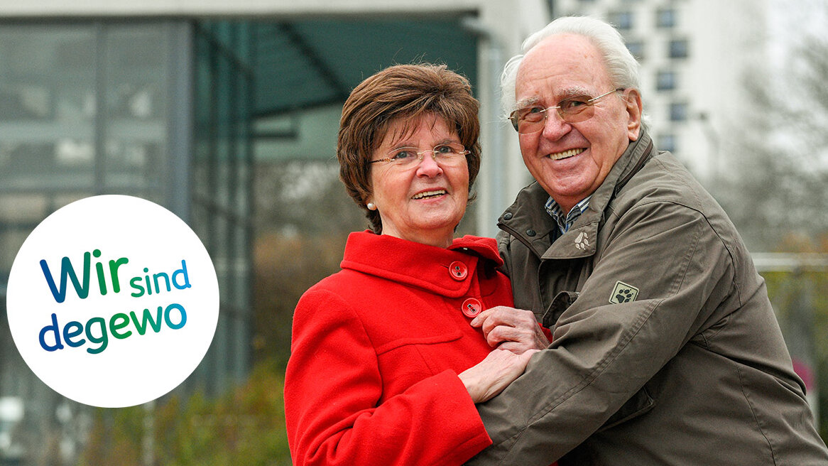 Bärbel und Udo Schulte in ihrer Nachbarschaft in Gropiusstadt. Udo Schulte trägt eine ockerfarbene Jacke und hat die Arme um seine Frau im roten Mantel gelegt. Beide lächeln in die Kamera.