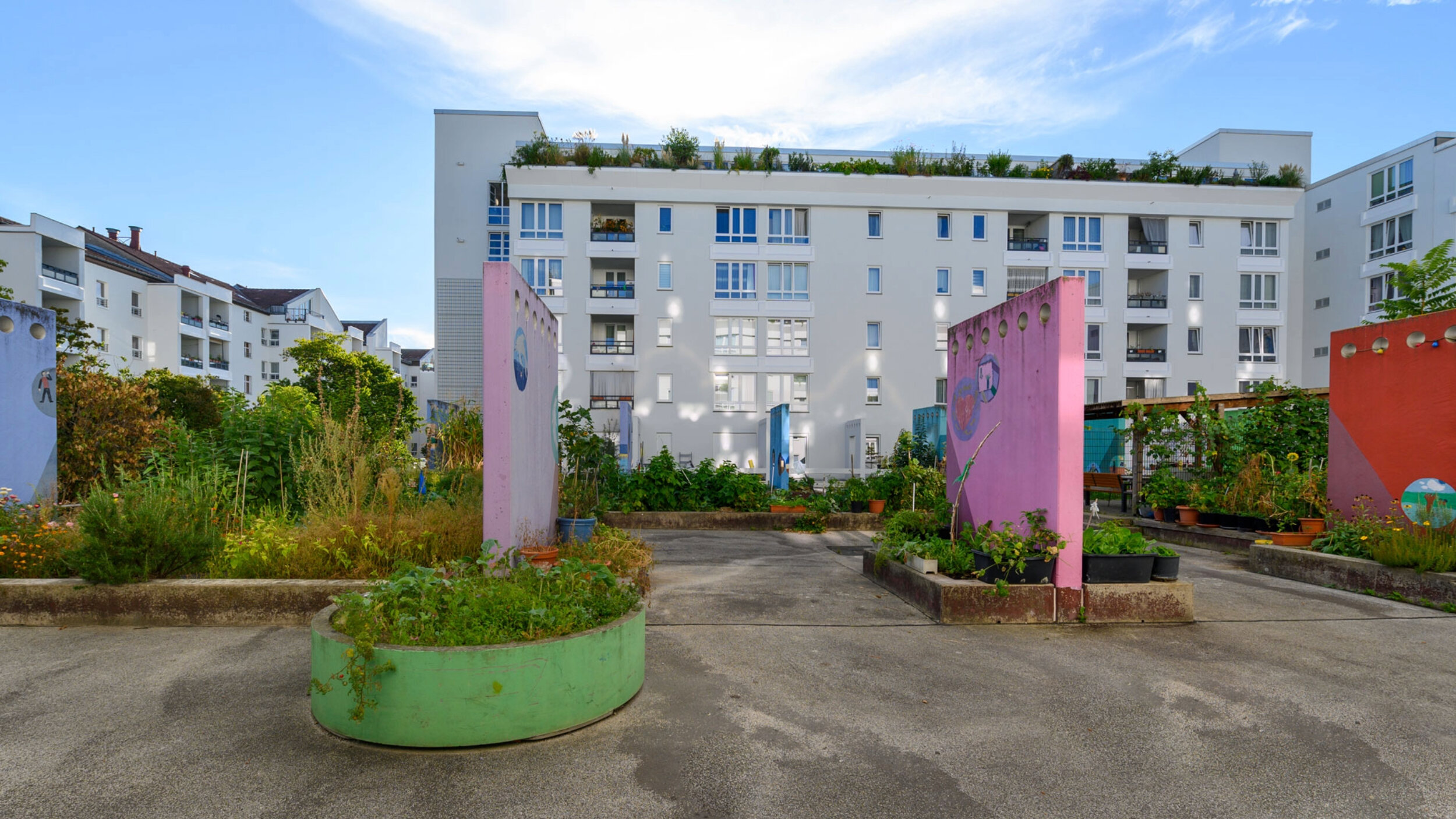 Innenhof einer Wohnanlage mit urbanem Gemeinschaftsgarten: bepflanzte Hochbeete und runde Pflanzinseln, farbige Gestaltungselemente in Rosa und Grün, im Hintergrund mehrgeschossige Wohngebäude mit Balkonen und Dachbegrünung.