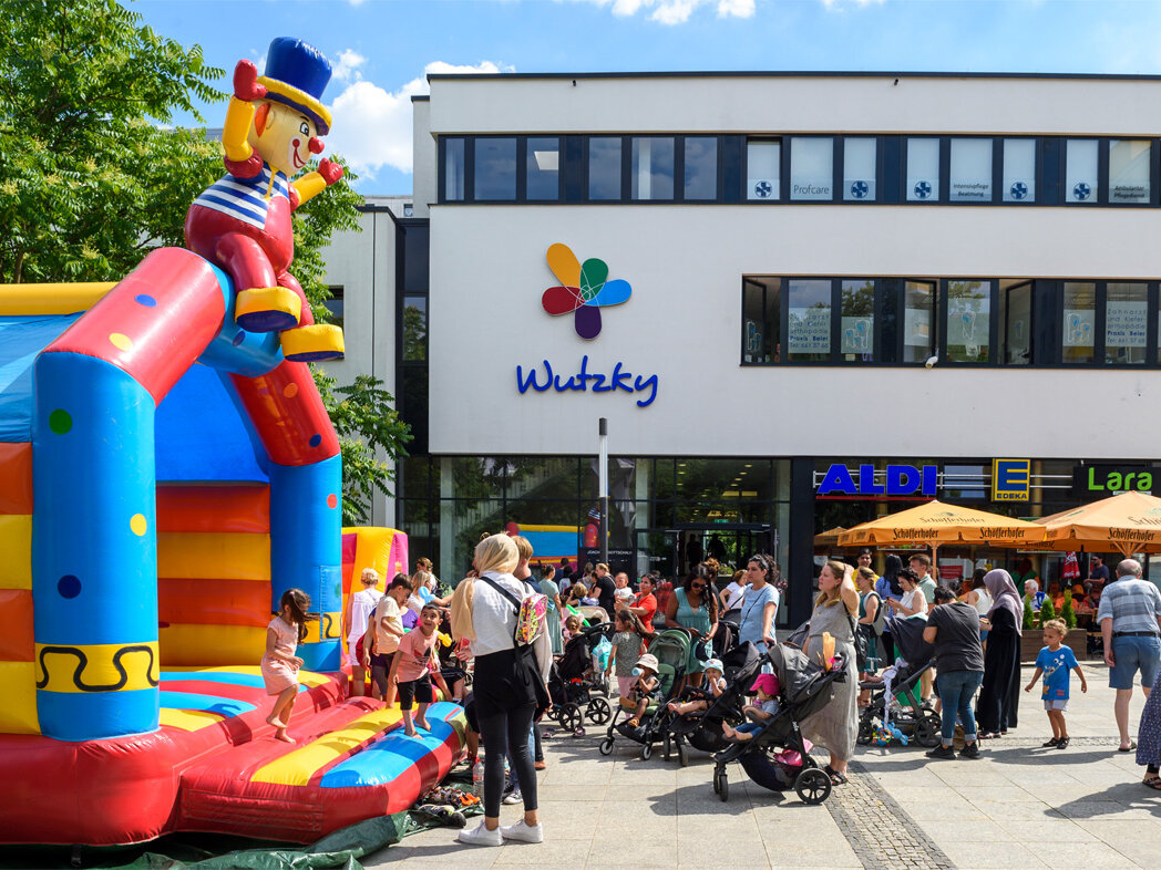 Menschen auf dem Sommerfest, links eine Hüpfburg, rechts gelbe große Sonnenschirme