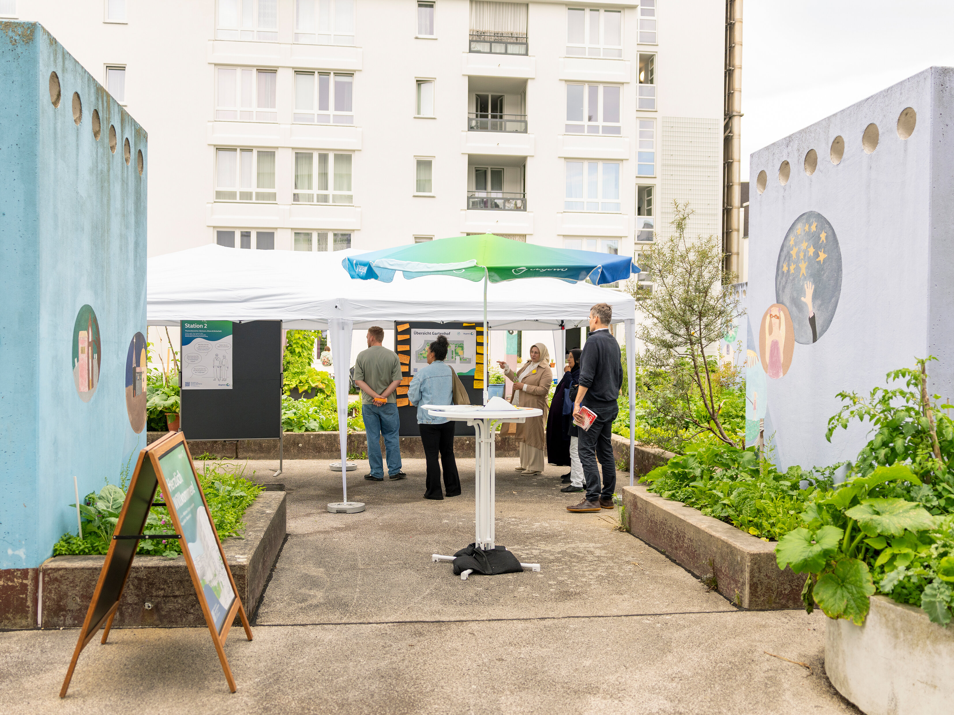 Übersicht über die Dialogstation im Innenhof: Pavillon, Informationswände und mehrere Teilnehmende zwischen Hochbeeten und Wohngebäuden.