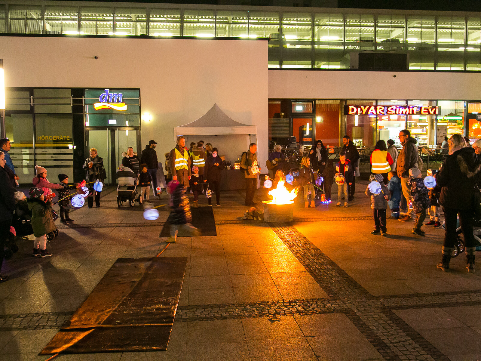 Ein Feuer mitten auf einem Platz, Menschen mit Laternen drum herum.
