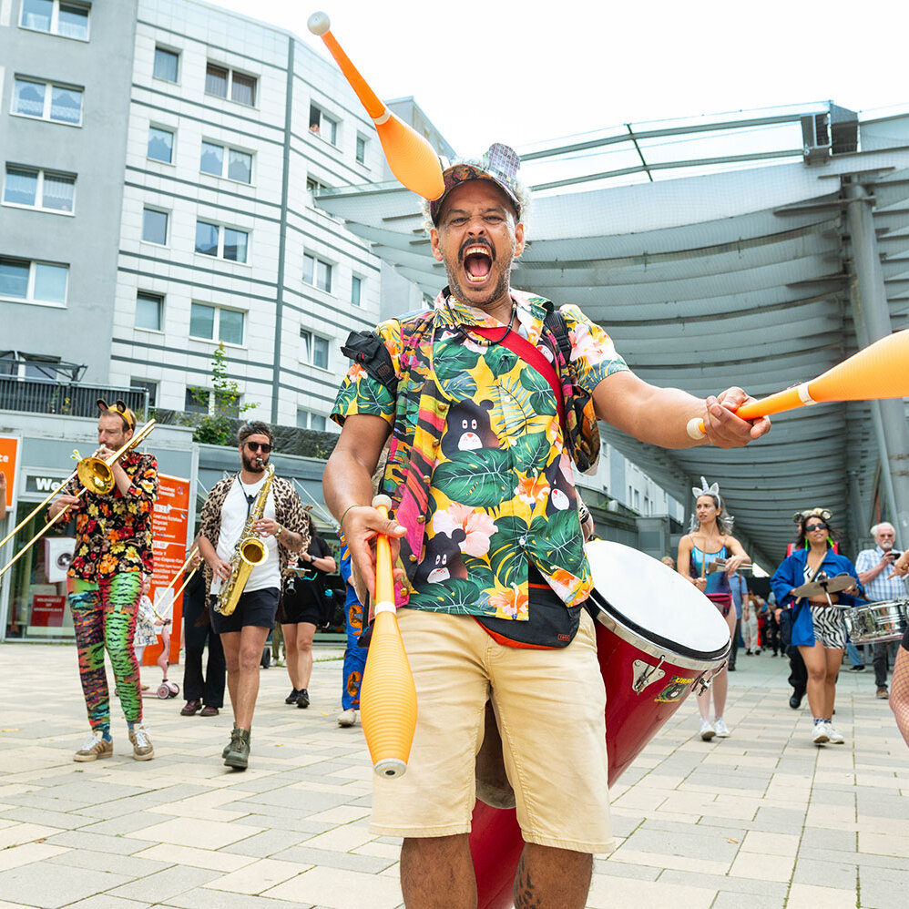 Ein Karnevalszug zieht auf der Marzahner Promenade entlang. Ein Mann jongliert mit Kegeln und lacht in die Kamera.