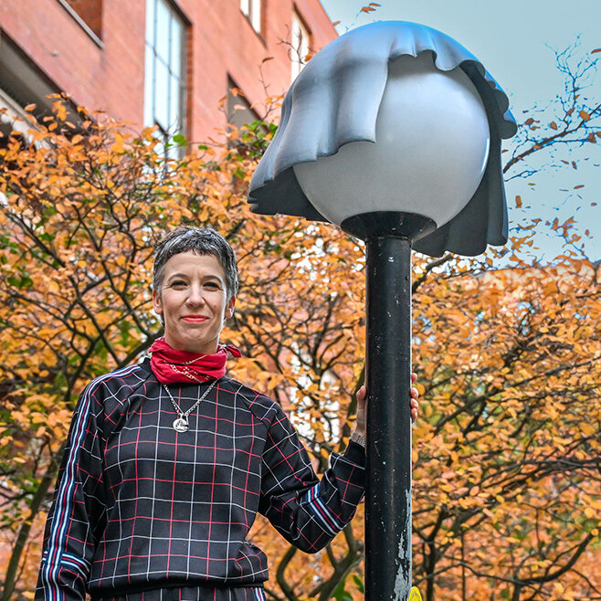 Frau steht an einer Laterne gelehnt. Die Laterne trägt eine Mütze.