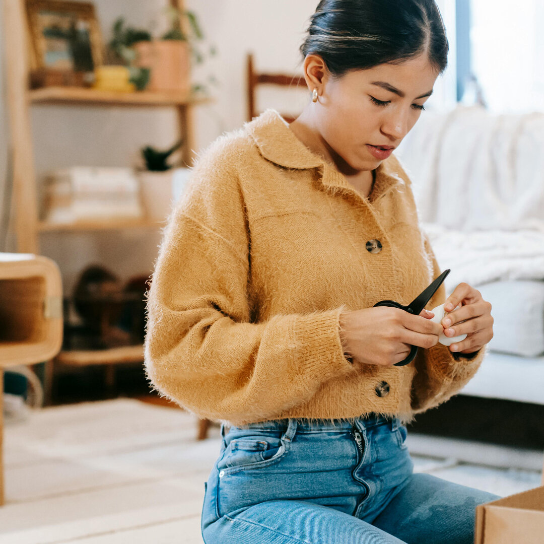 Person sitzt in einer Wohnung auf dem Boden und öffnet mit einer Schere einen Karton. Im Hintergrund sind Sofa, Regal und Wohnaccessoires zu sehen.