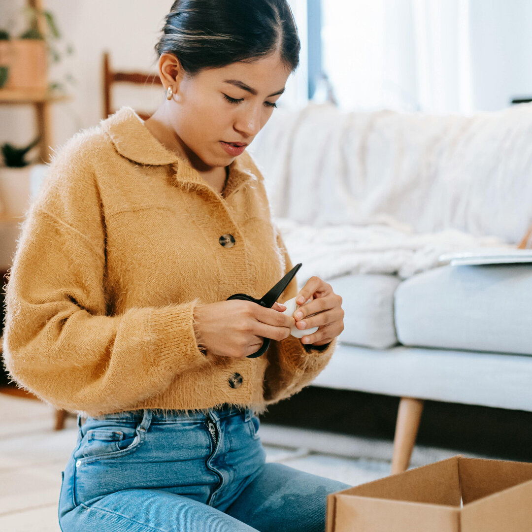 Person sitzt in einer Wohnung auf dem Boden und öffnet mit einer Schere einen Karton. Im Hintergrund sind Sofa, Regal und Wohnaccessoires zu sehen.