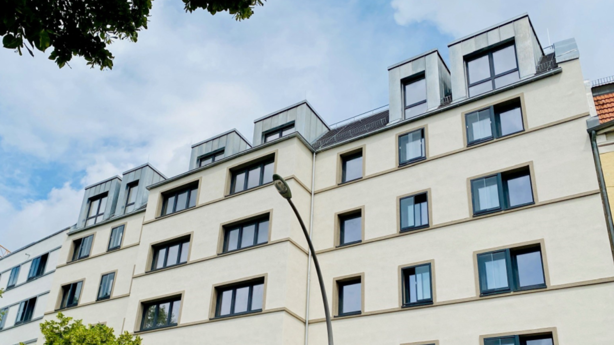 Mehrgeschossiges Wohngebäude mit heller Putzfassade und regelmäßig angeordneten Fenstern. Das Dach ist als ausgebautes Dachgeschoss mit Gauben ausgeführt. Straßenlaterne und Bäume befinden sich im Vordergrund.