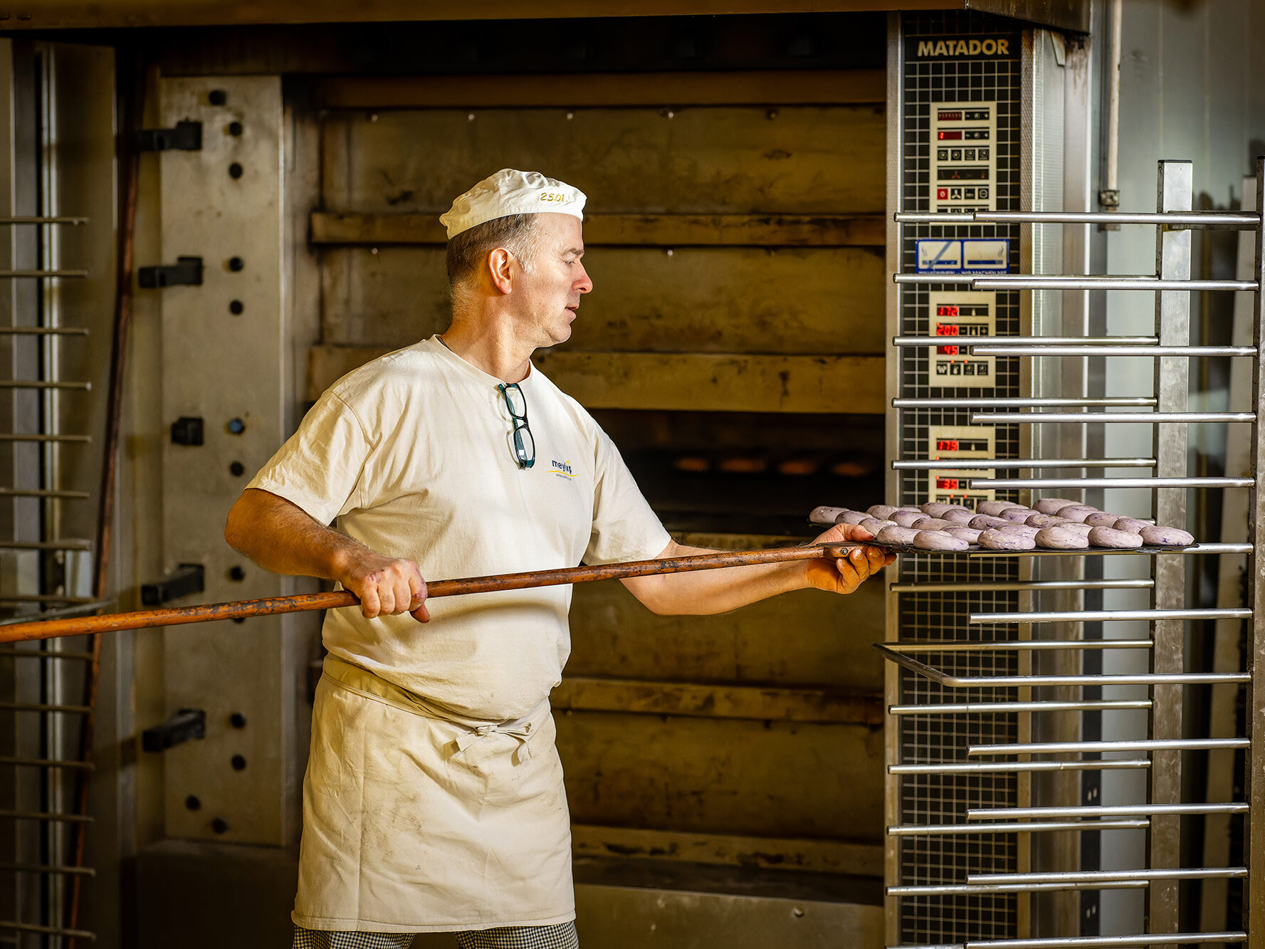 Bäcker schiebt mit einer langen Holzschaufel ein Blech mit rohen Teiglingen vor den großen Backofen.