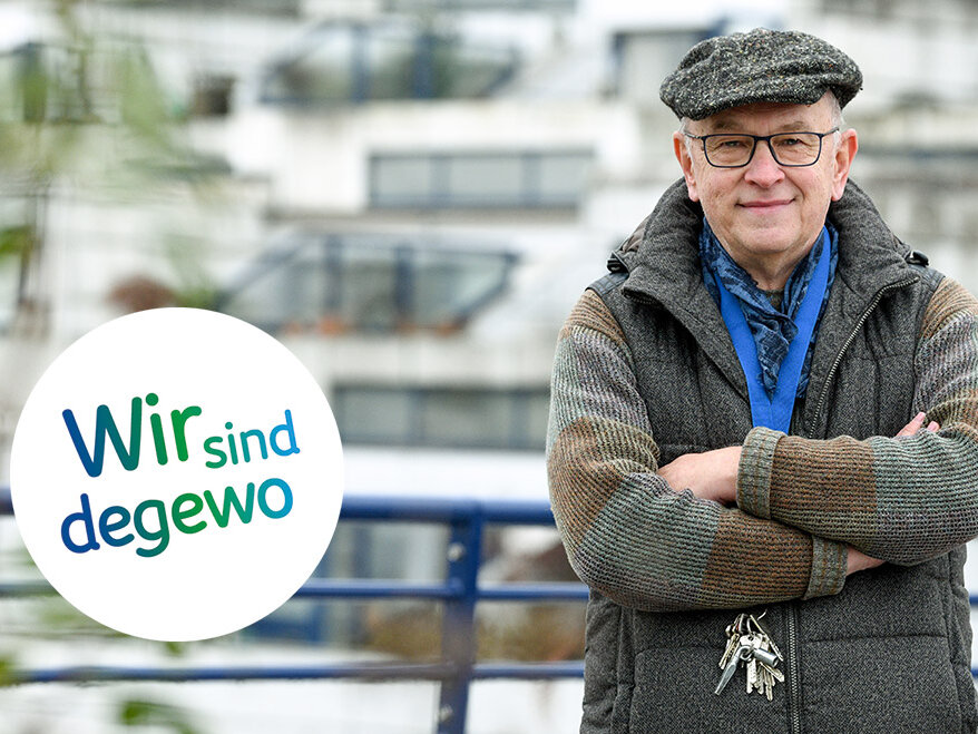 Ein älterer Mann mit Mütze und Brille steht mit verschränkten Armen im Freien vor einem Wohngebäude während das degewo Logo mit der Aufschrift Wir sind degewo eingeblendet ist.