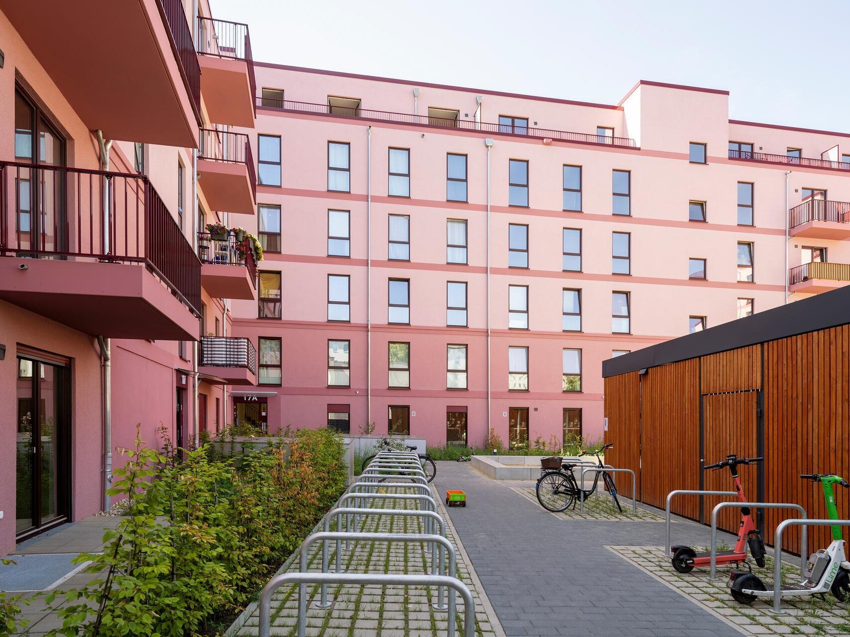 Neubau Bismarckstraße in Spandau, Straßenansicht des Mehrfamilienhauses mit roter Fassade, Staffelgeschoss und Balkonen. Vor dem Gebäude verlaufen Gehweg und Fahrbahn.