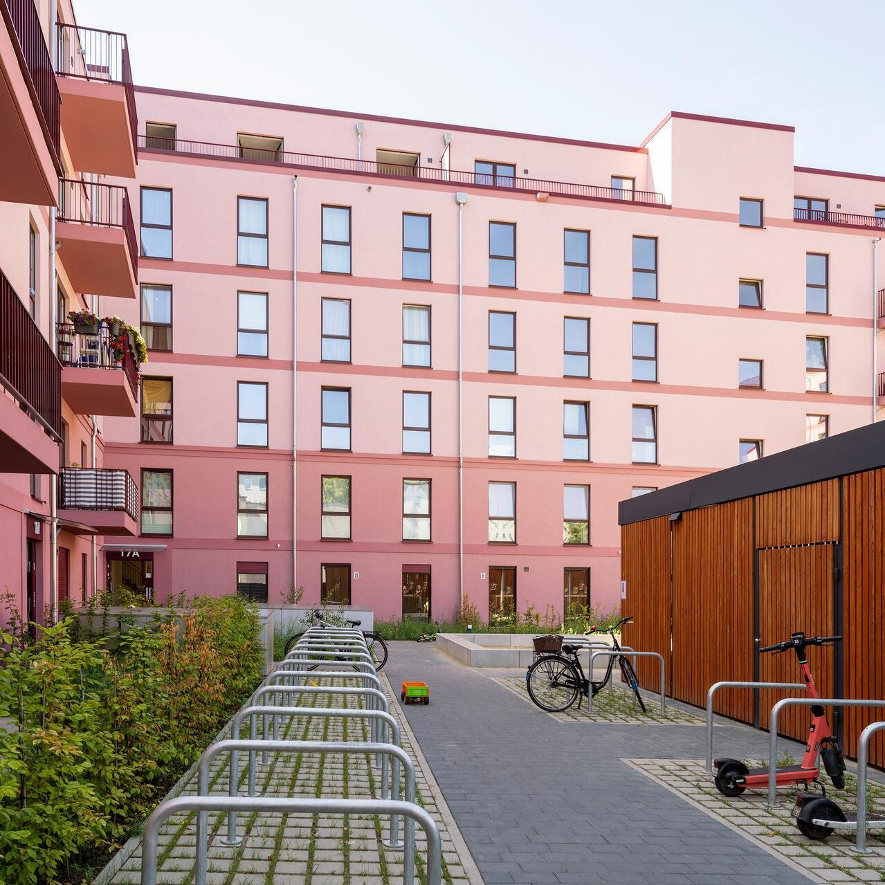Neubau Bismarckstraße in Spandau, Straßenansicht des Mehrfamilienhauses mit roter Fassade, Staffelgeschoss und Balkonen. Vor dem Gebäude verlaufen Gehweg und Fahrbahn.