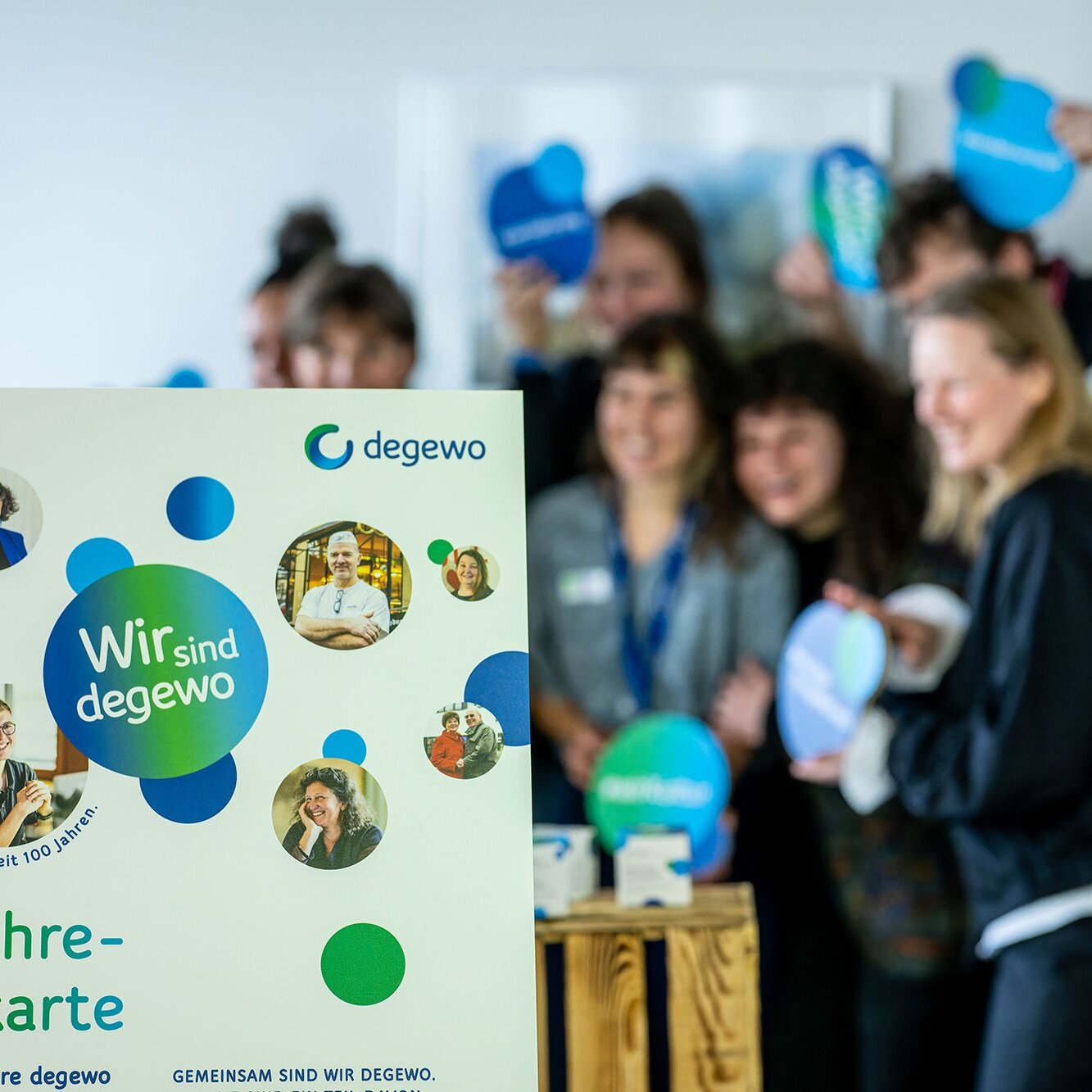 Im Vordergrund steht ein Aufsteller mit der Aufschrift „Wir sind degewo“ und „100-Jahre-Berlinkarte“ sowie mehreren Porträtfotos. Im Hintergrund posiert eine Gruppe von Personen mit runden Schildern in Blau- und Grüntönen. Die Personen sind unscharf dargestellt, während der Aufsteller im Fokus steht.